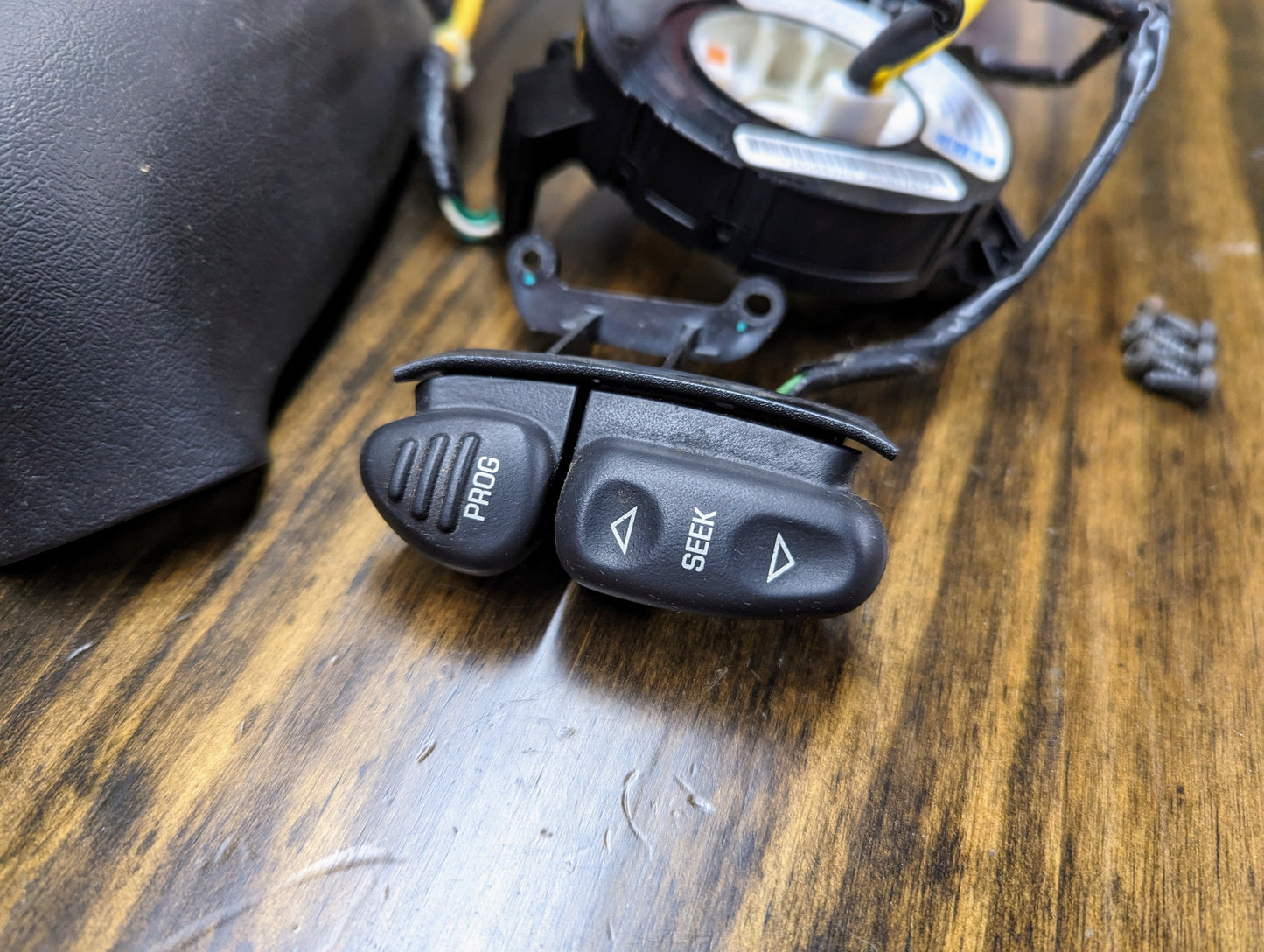 Close-up of General Motors KIT Steering Wheel Radio Controls & Clock Spring for 1995-2002 GM Full Size OBS/NBS Trucks, showing black PROG and SEEK buttons on wood with wiring and car components.