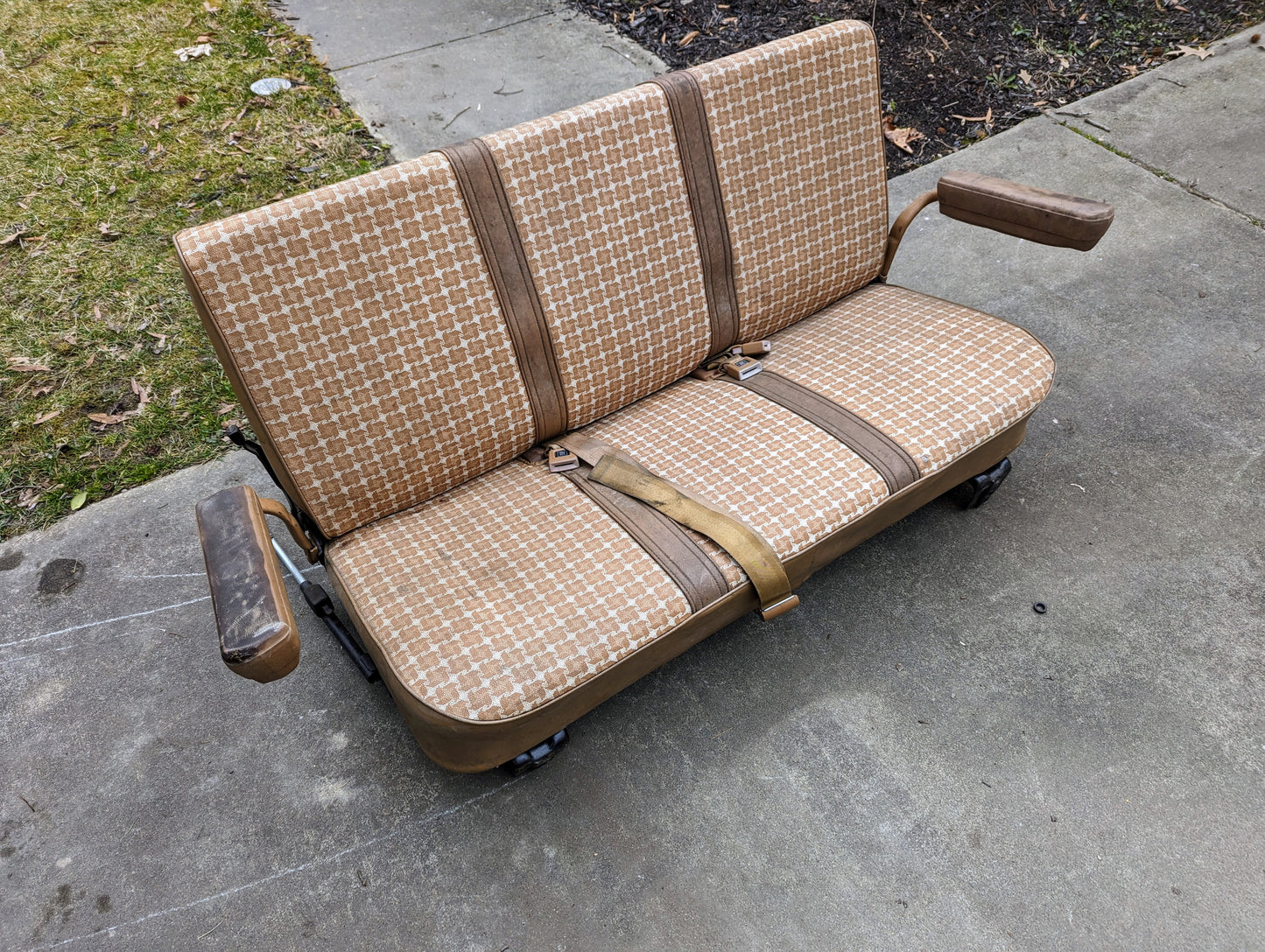A General Motors OEM Rear Seat in Tan Pattern for 1973–1991 K5 Blazer & Jimmy, with seat belts, padded armrests, and sturdy mounts, sits on a concrete driveway near a grassy area with fallen leaves.