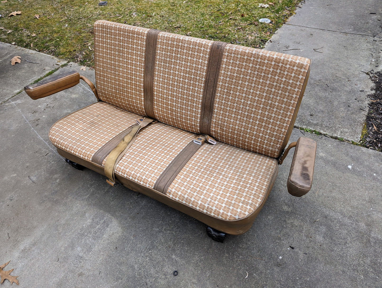 A General Motors OEM Rear Seat in Tan Pattern for 1973-1991 K5 Blazer & Jimmy, featuring seat belts, armrests, and mounts, sits on a sidewalk next to grass and pavement.