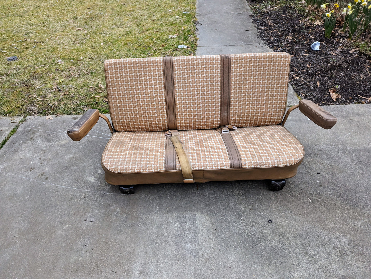 A General Motors OEM Rear Seat in tan pattern for 1973–1991 K5 Blazer & Jimmy, featuring seat belts, attached armrests, and original mounts, sits on a driveway—perfect for your classic restoration project.