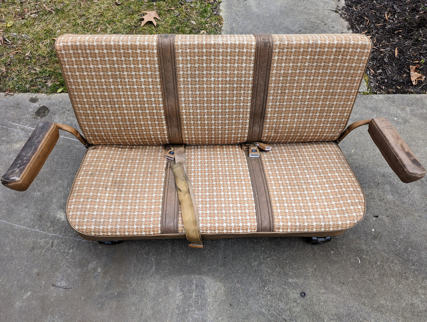 A General Motors OEM Rear Seat in tan pattern for 1973–1991 K5 Blazer & Jimmy, with seat belts and worn armrests, sits outdoors on concrete. Seat mounts are visible, with dry leaves and grass scattered in the background.