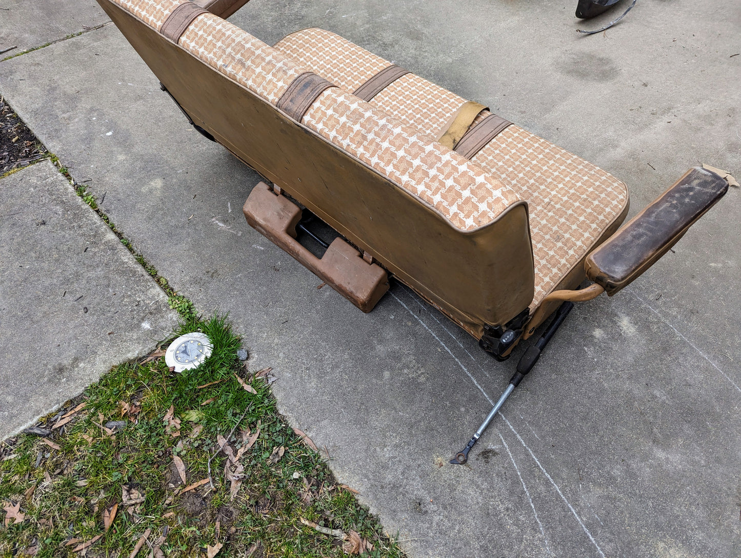 A General Motors OEM Rear Seat in Tan Pattern for 1973-1991 K5 Blazer & Jimmy sits on a concrete driveway near grass, with seat mounts and hardware visible underneath. A round metal object is embedded in the ground nearby.