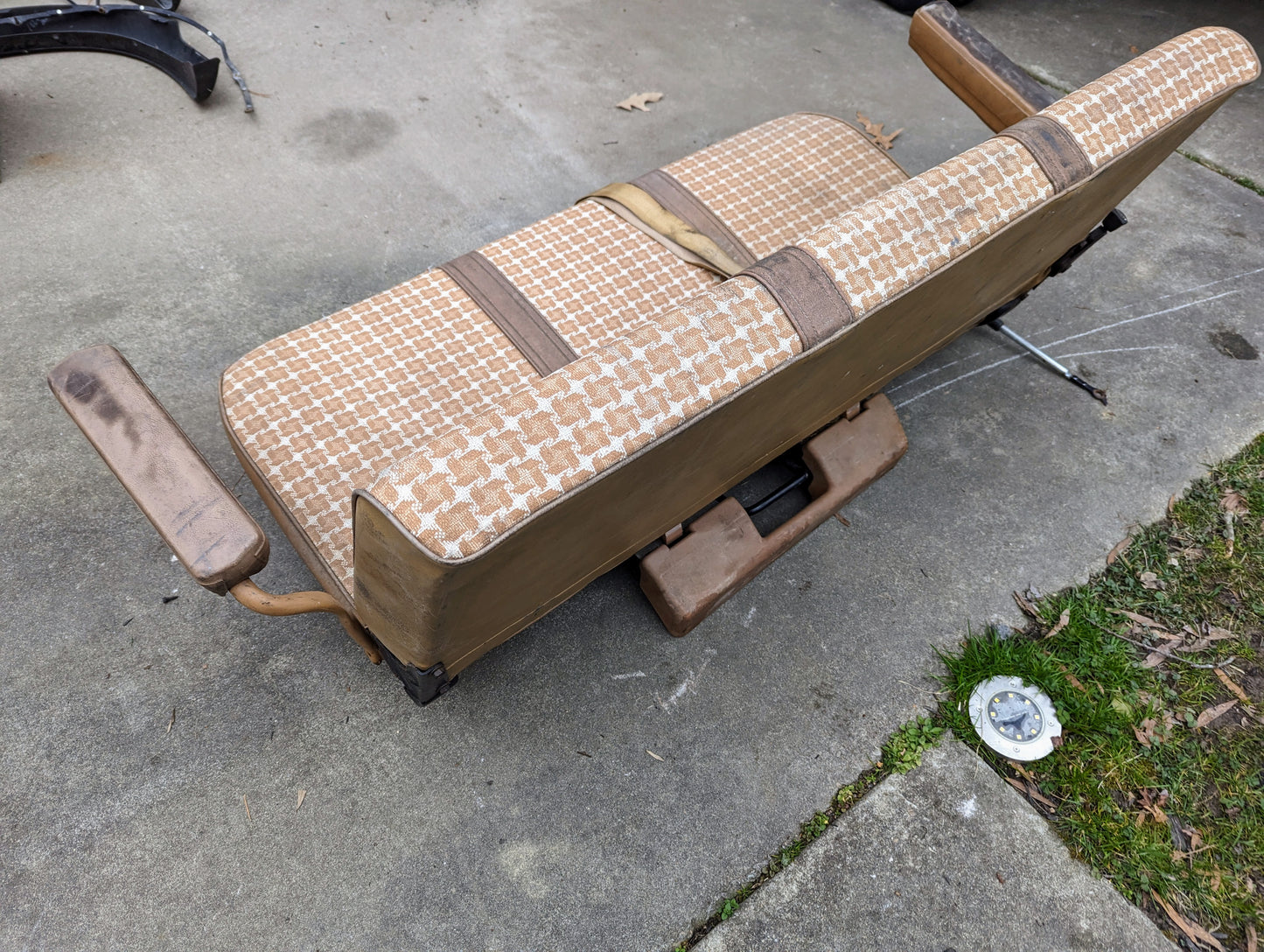 A General Motors OEM Rear Seat in tan pattern for 1973-1991 K5 Blazer & Jimmy rests on a concrete driveway near grass, featuring original seat mounts and displaying age, wear, and signature vintage style.