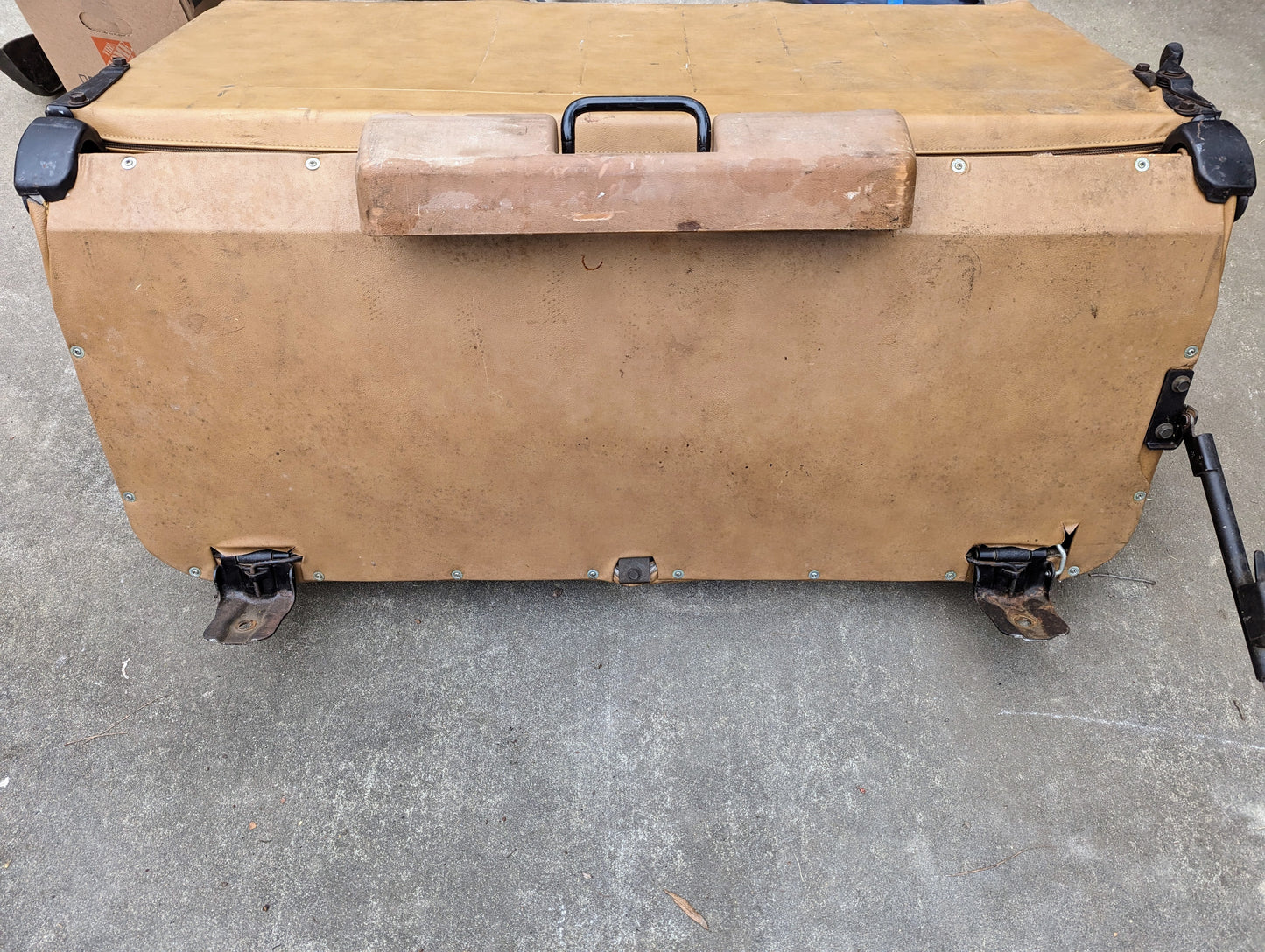 A tan OEM Rear Seat in Tan Pattern for 1973-1991 K5 Blazer & Jimmy by General Motors lies flat on concrete near seat mounts. It shows signs of use, with visible scuffs and dirt on its surface.