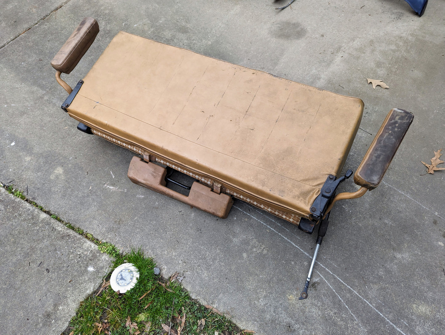 A General Motors OEM rear seat in tan pattern for 1973-1991 K5 Blazer & Jimmy sits on a concrete driveway near grass and scattered leaves, its padded armrests, metal frame, and visible seat mounts easily seen underneath.