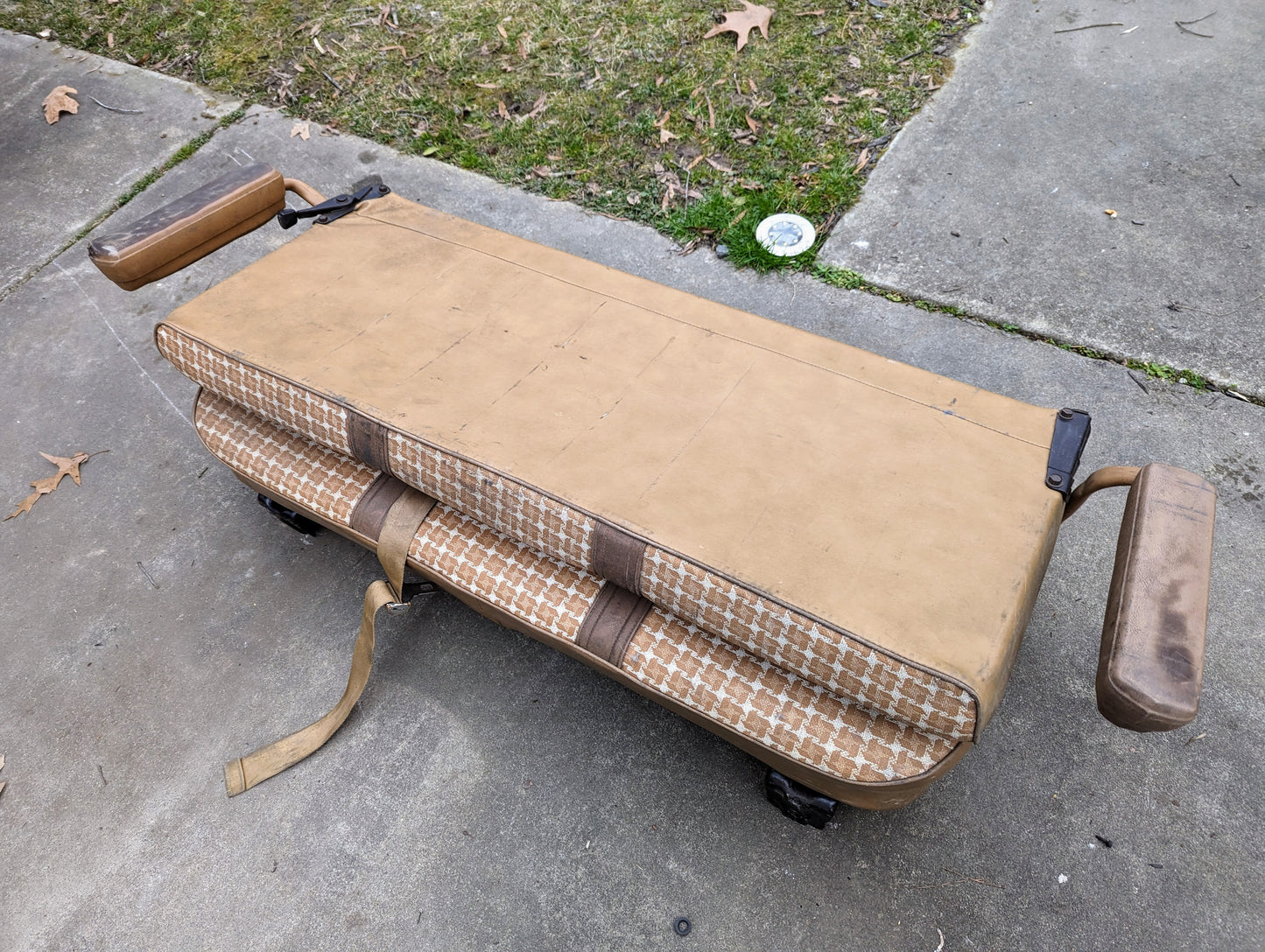 A General Motors OEM Rear Seat in Tan Pattern for 1973-1991 K5 Blazer & Jimmy, with seat mounts visible, sits outdoors on concrete amid grass and leaves.