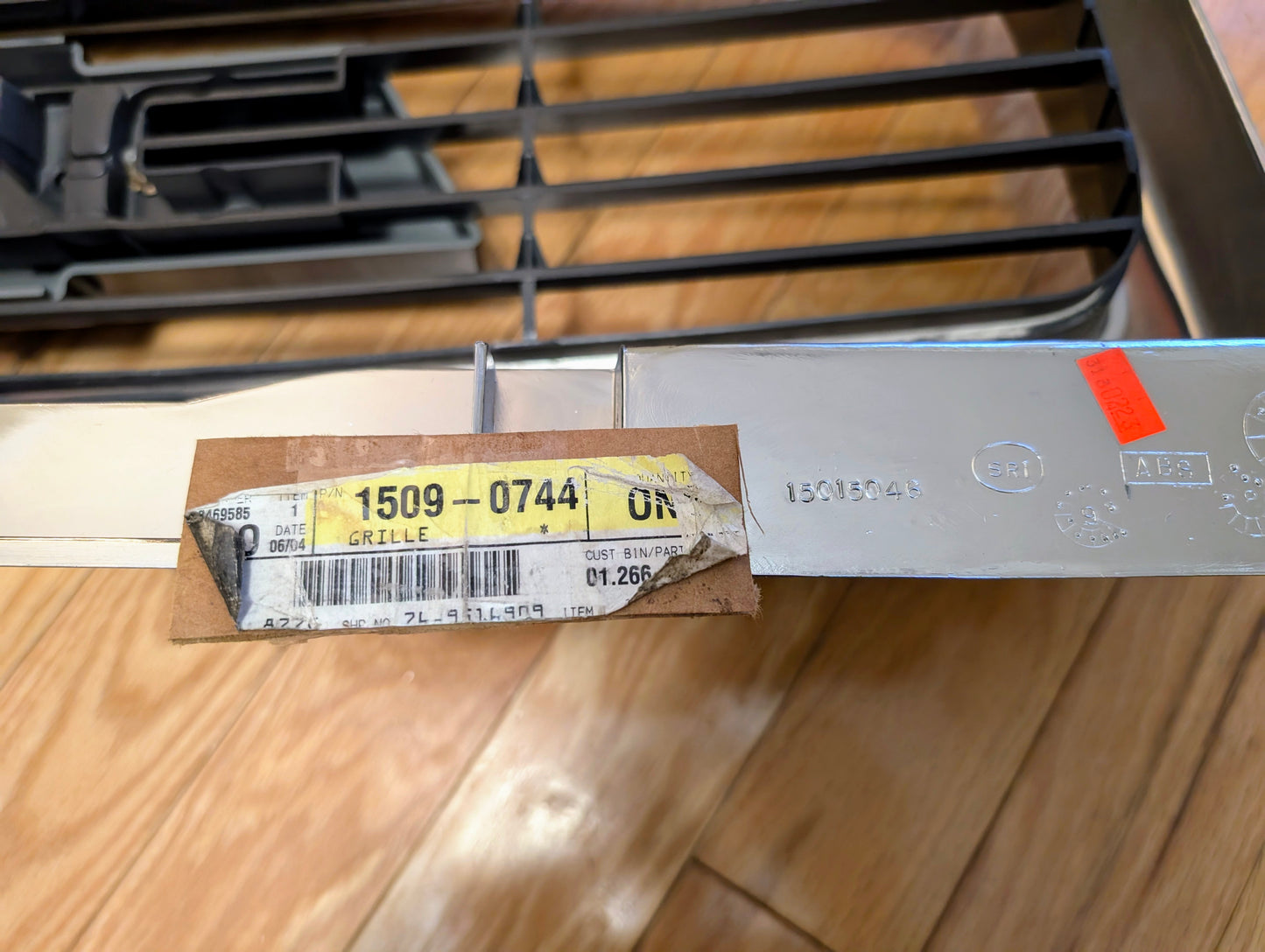 A close-up of a metal car part with a sticker labeled 1509-0744 GRILLE and a barcode, resting on a wood floor. An OEM General Motors NOS Export Front Radiator Grille in chrome for 1998-2005 Chevrolet S10, Blazer, and more is partially visible in the background.