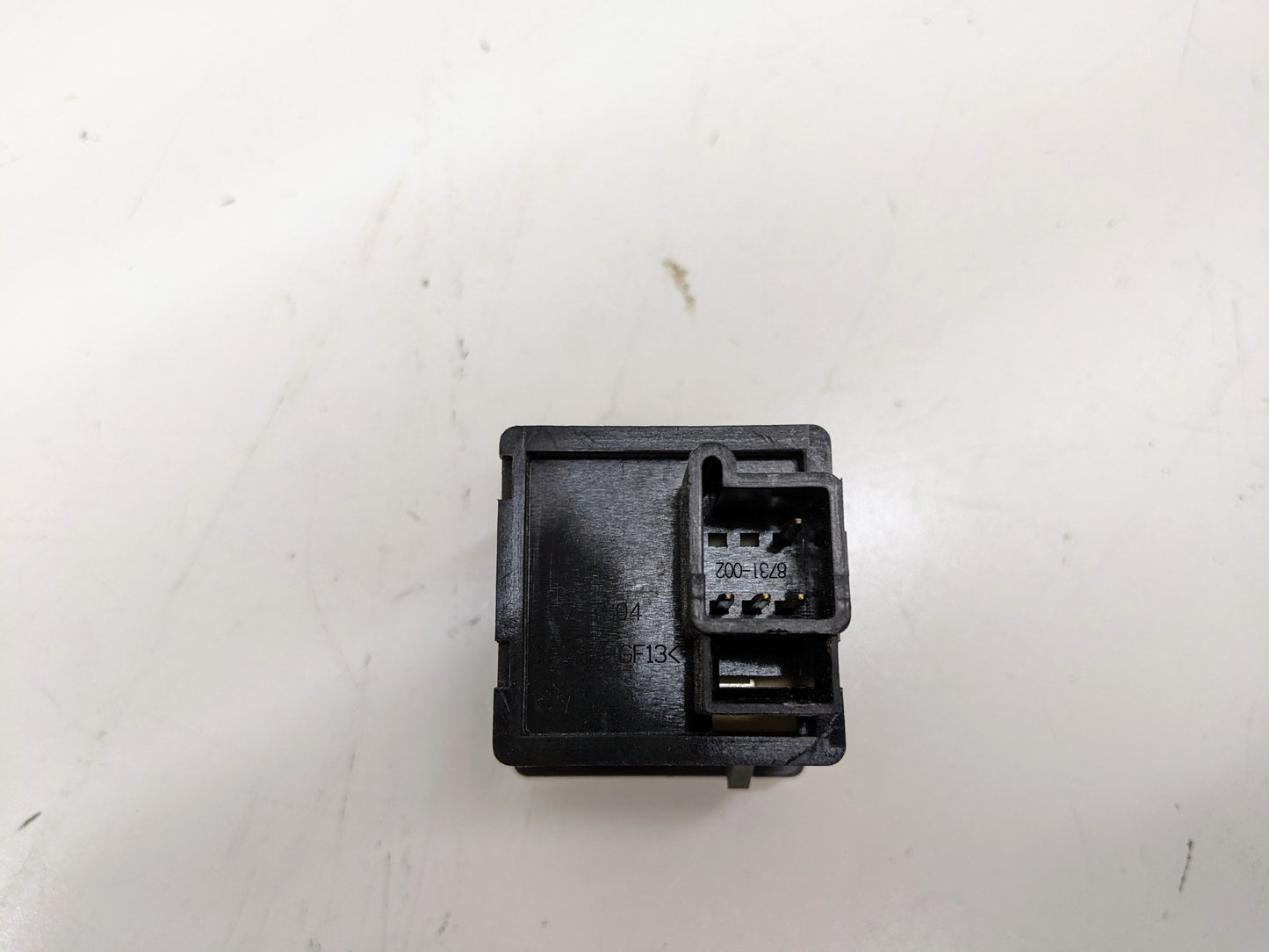 A General Motors 2003-2006 Escalade, Tahoe, Yukon, Suburban, Avalanche, Savana Traction Control Switch with metal prongs and connector port rests on a slightly dirty white surface; part numbers are visible on its top.