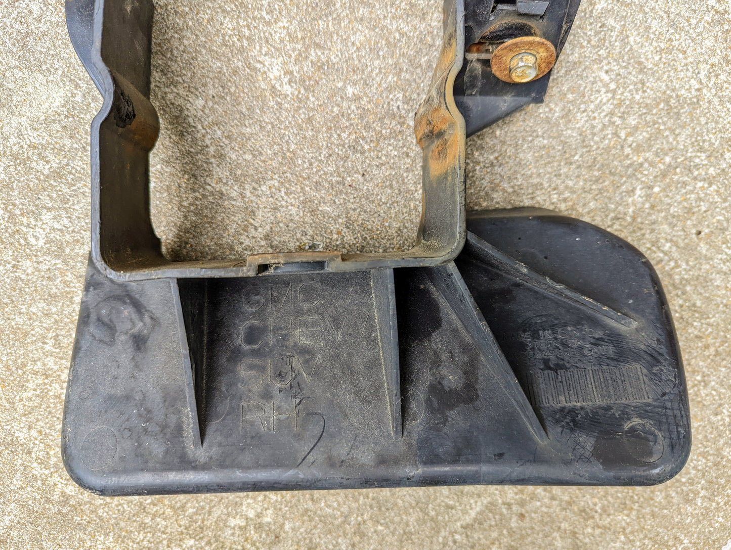 A black plastic General Motors Bumper Deflector Tow Hook Aero Panel for 2003-2006 Chevrolet Tahoe rests on rough concrete, showing a rusted bolt and visible wear—suitable for Tahoe and similar GMC Yukon models.