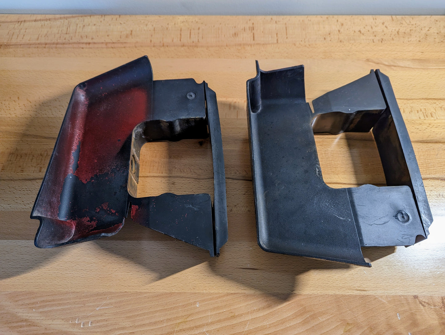 Two General Motors Bumper Deflector Tow Hook Aero Panels for 2003-2006 GMC Sierra 1500 are upright on a wooden surface—left panel has chipped red paint, right one shows an even black finish.