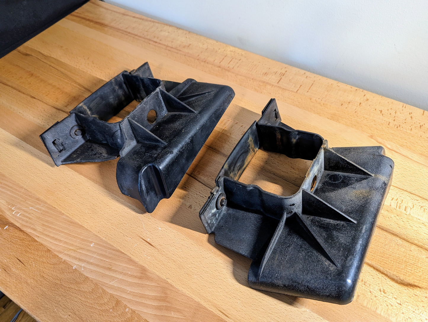 Two black plastic General Motors Bumper Deflector Tow Hook Aero Panels for 2003-2006 GMC Sierra 1500, showing dust and wear near the mounting holes, rest on a wooden workbench.