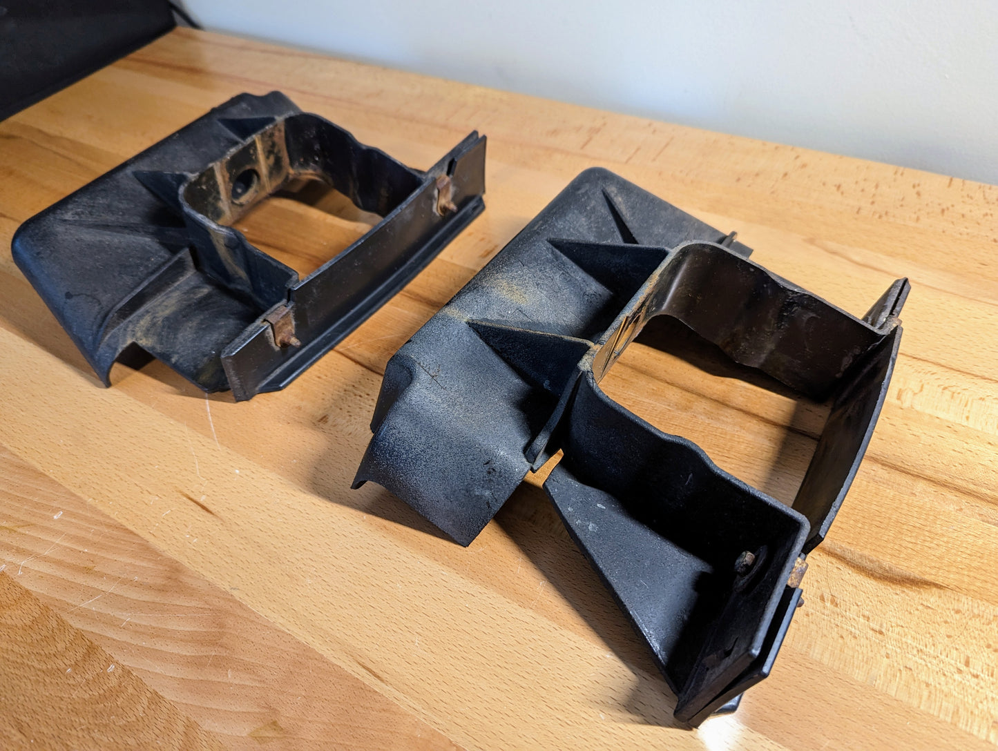 Two black General Motors Bumper Deflector Tow Hook Aero Panels for 2003-2006 GMC Sierra 1500, featuring rectangular openings and angled edges, are arranged parallel on a wooden surface, showing minor dust and wear.