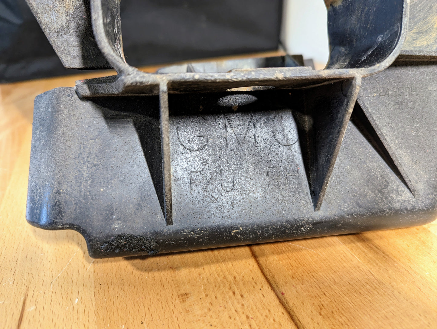 A close-up of a dusty black General Motors Bumper Deflector Tow Hook Aero Panel for 2003-2006 GMC Sierra 1500, with GM lettering and faint text visible on its surface, resting on a wooden surface.