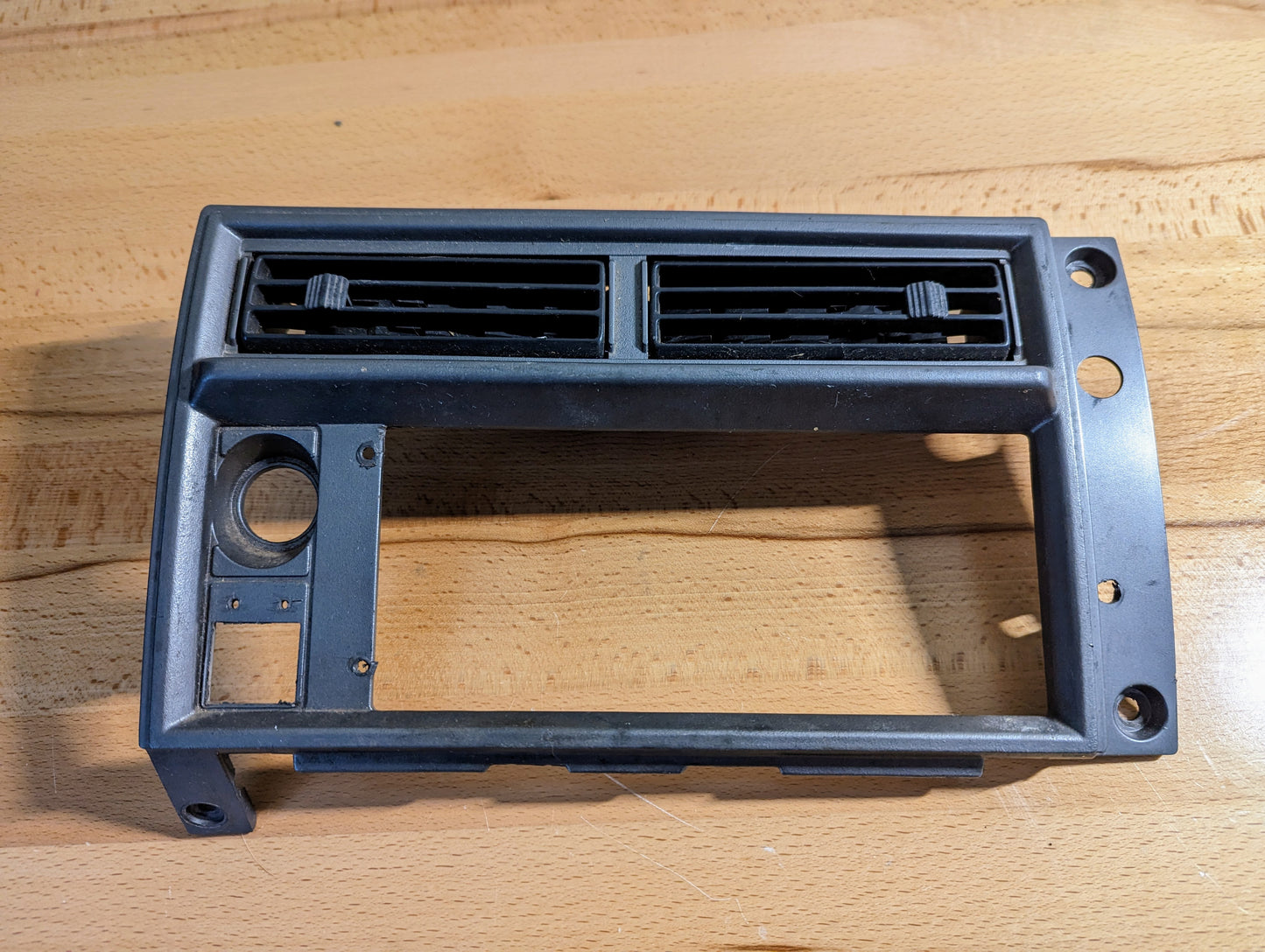 A silver OEM Radio Dash Bezel by General Motors for 1986-1993 Chevy S10, Blazer, and GMC S15, with two horizontal air vents and slots, shown on a wooden surface.