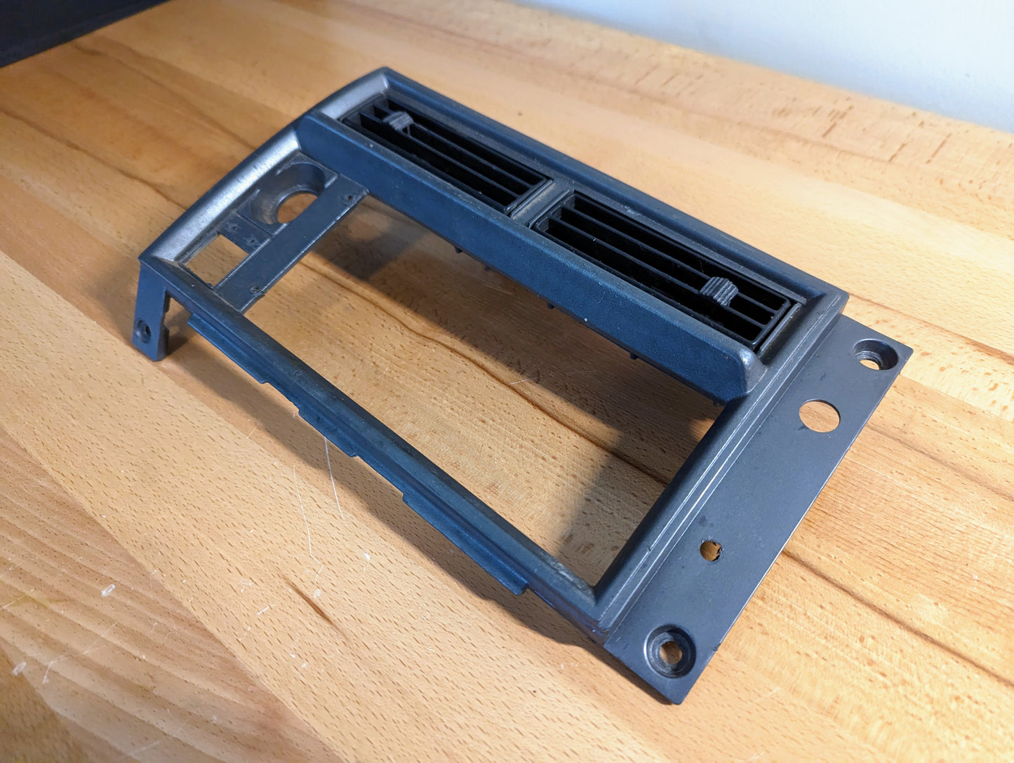 A silver OEM Radio Dash Bezel by General Motors for 1986-1993 Chevy S10, Blazer, and GMC S15 features two rectangular air vents, mounting holes, and cutouts for extra controls. The trim rests on a light wood surface.