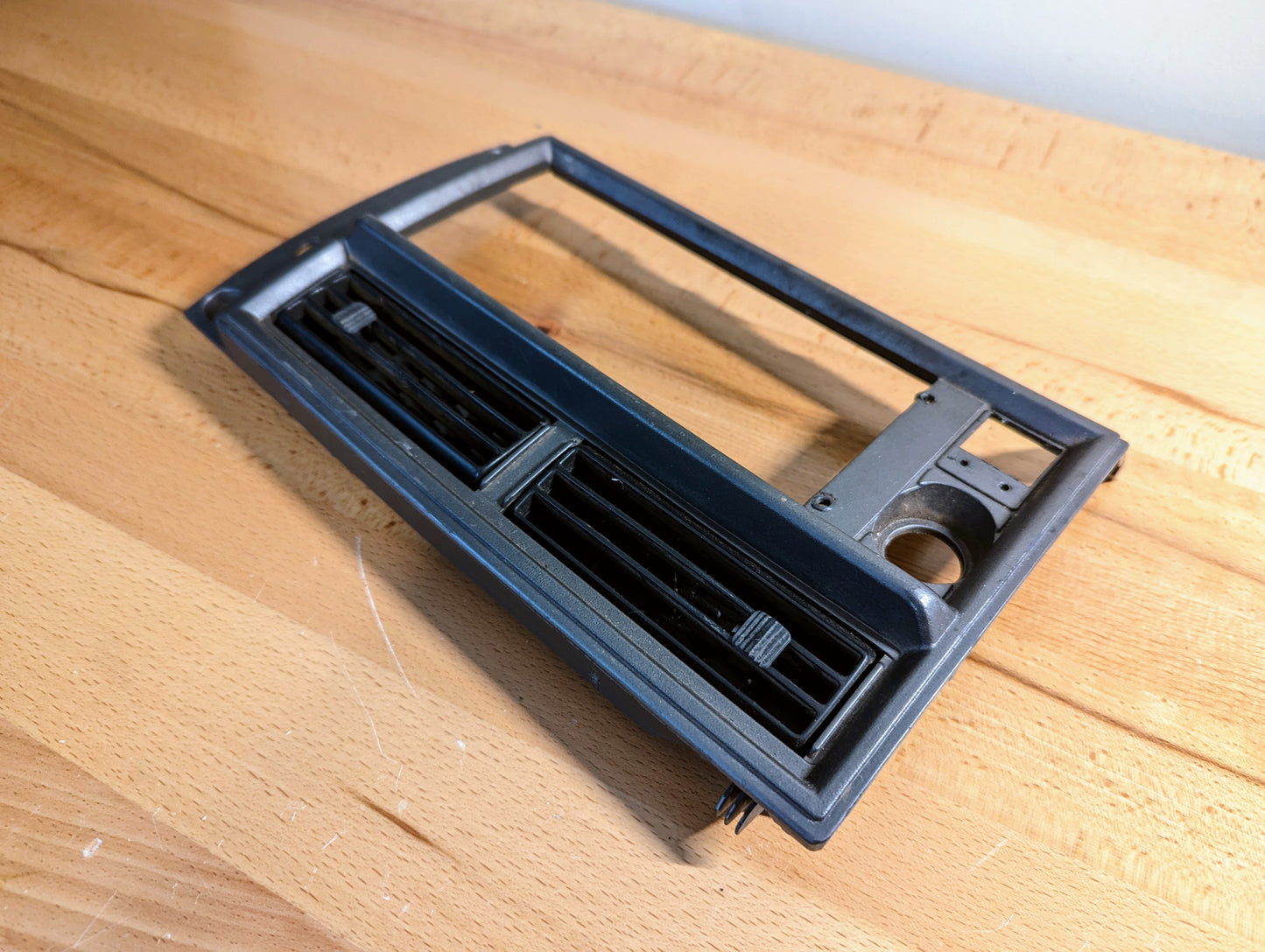 A silver OEM Radio Dash Bezel by General Motors for 1986-1993 Chevy S10 Blazer and GMC S15, designed with air vents and a radio slot, is displayed on a wooden surface.