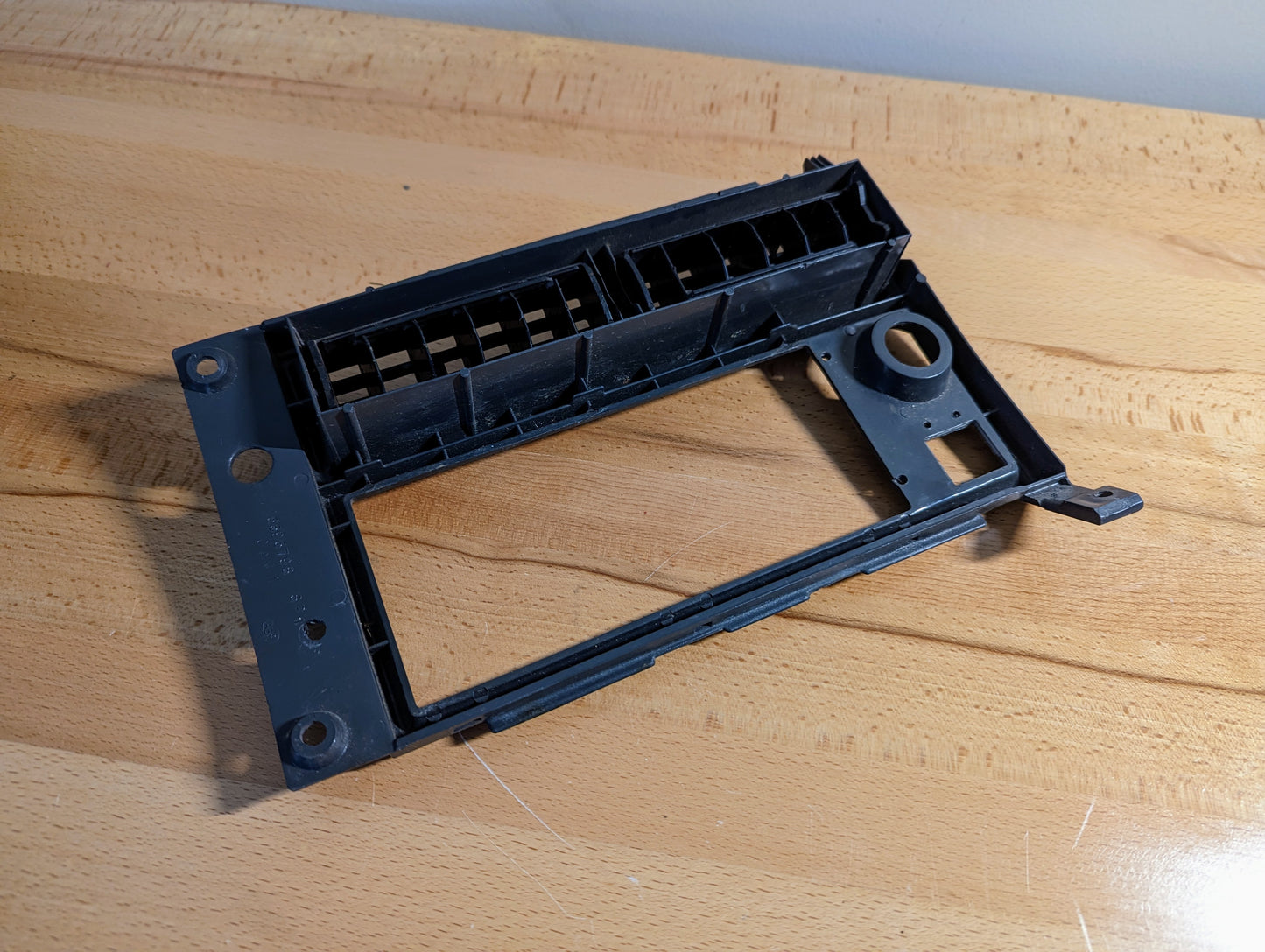 A silver General Motors OEM Radio Dash Bezel for 1986-1993 Chevy S10, Blazer, GMC S15, and more rests on a light wooden surface, displaying slots and openings for audio equipment.