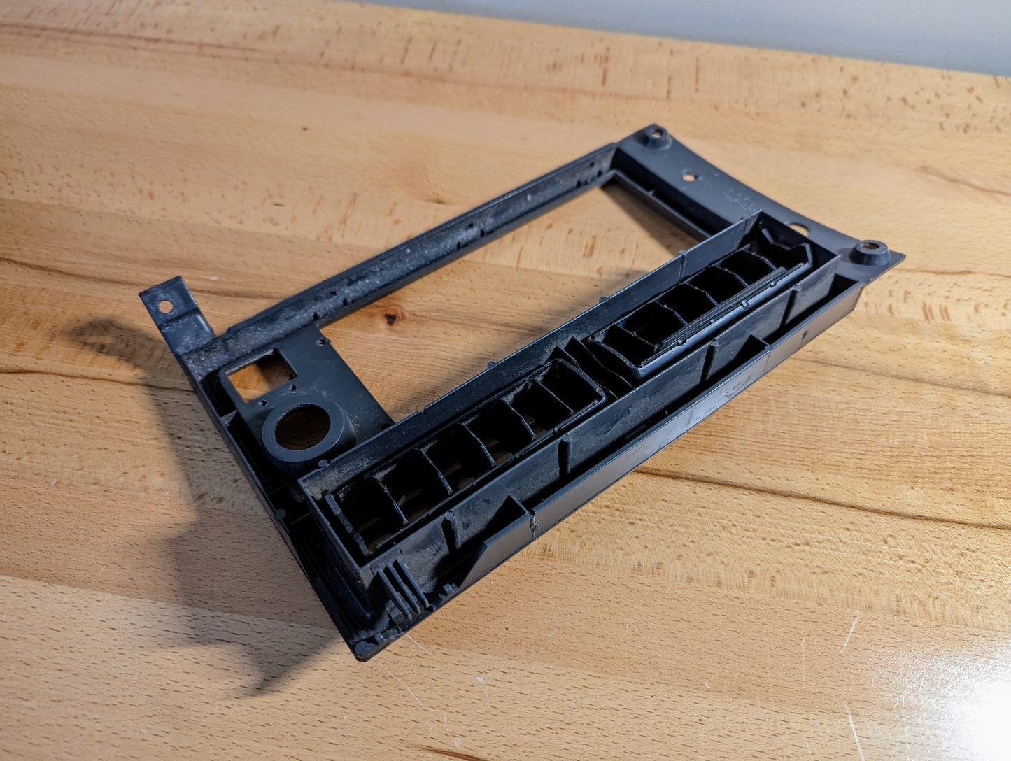 A silver General Motors OEM Radio Dash Bezel for 1986-1993 Chevy S10, Blazer, and GMC S15 rests on a light wood surface. The trim has multiple mounting points and cutouts.