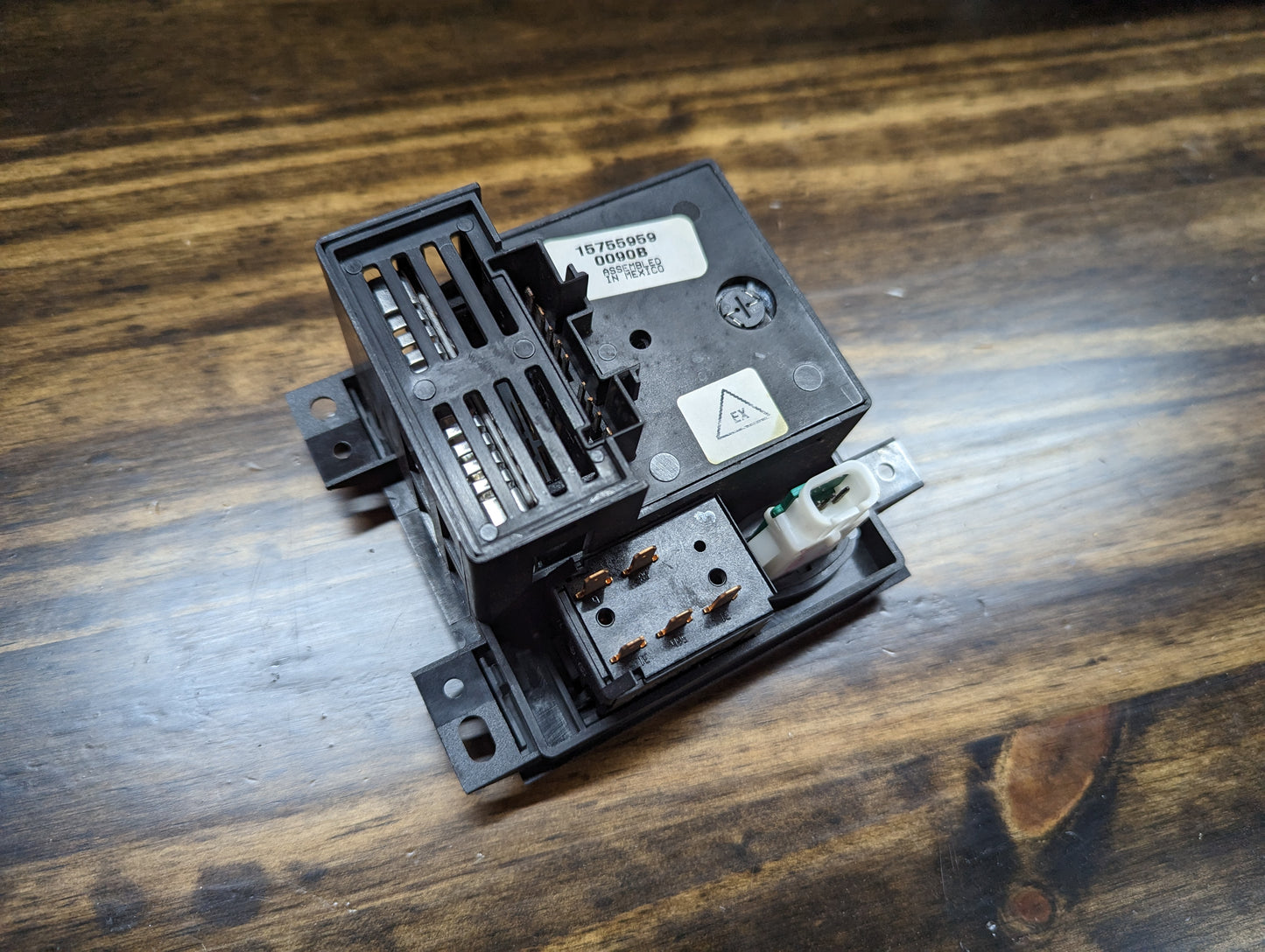 A General Motors NOS Headlight Switch with rear fog and level adjust for 1998-2005 Chevy S10, Blazer, GMC Sonoma, and Jimmy rests on a wooden surface, showing exposed connectors, a white plug socket, and labeled top.