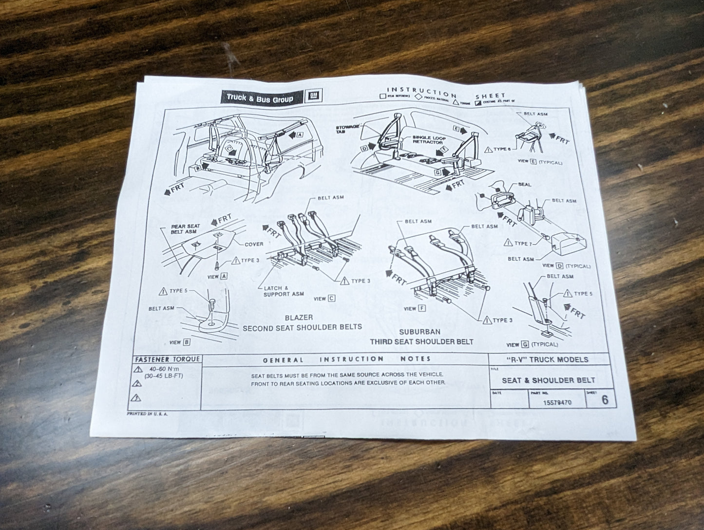 Instruction sheet on a wooden table shows diagrams and labeled parts for installing the General Motors NOS Passenger Seat Belt Retractor in Blue for 1973-1987 Chevy/GMC C10/K10 trucks, with detailed assembly steps and notes.