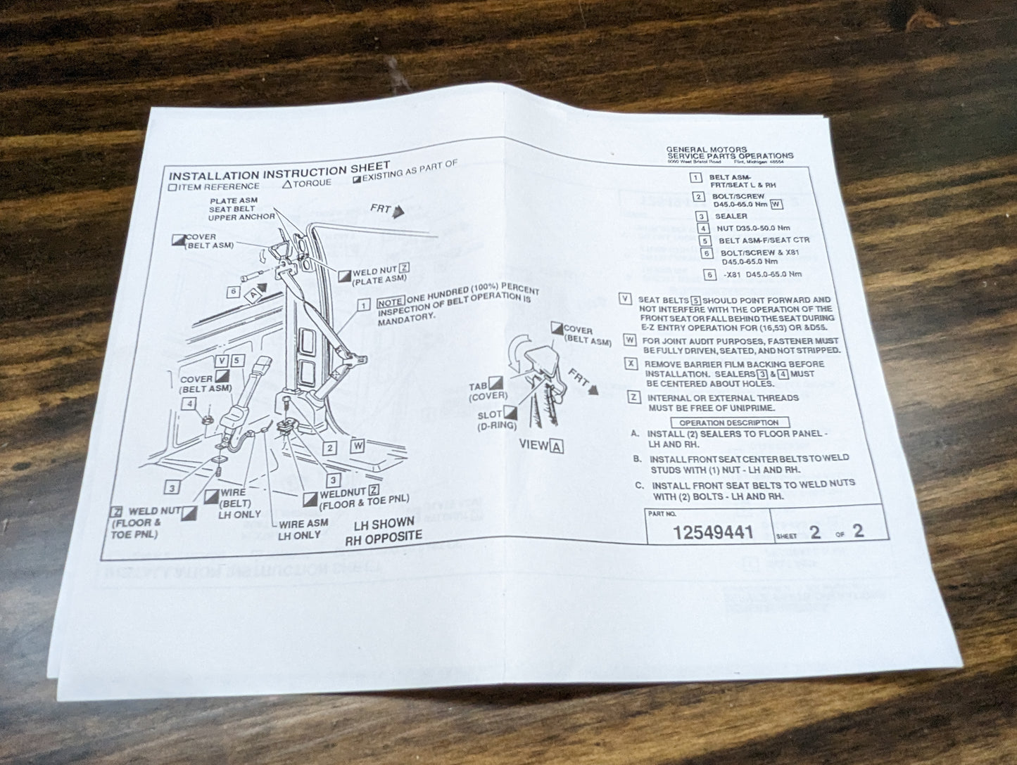 A printed instruction sheet with diagrams and labeled parts for the General Motors NOS Passenger Seat Belt Retractor in Red (1988–1998 Chevy/GMC C/K Trucks) is laid flat on a wooden surface, featuring step-by-step illustrations and a parts list.
