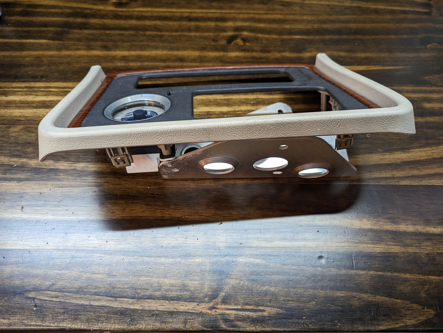 A General Motors OEM Center Console Front Bin Woodgrain Trim with Clock for the 2002 Cadillac Escalade, featuring beige and brown accents, is shown on a wooden surface with visible cutouts for controls and internal components.