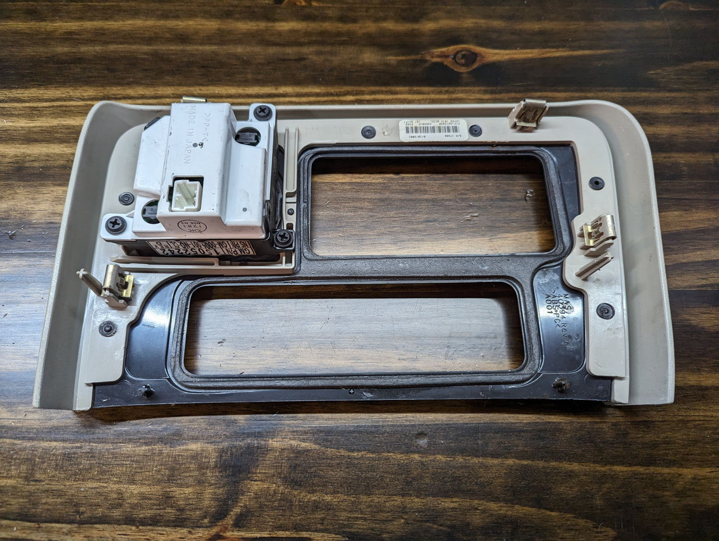 A General Motors OEM Center Console Front Bin Woodgrain Trim with Clock for a 2002 Cadillac Escalade sits on a wooden surface, featuring a beige and black finish with two large openings.