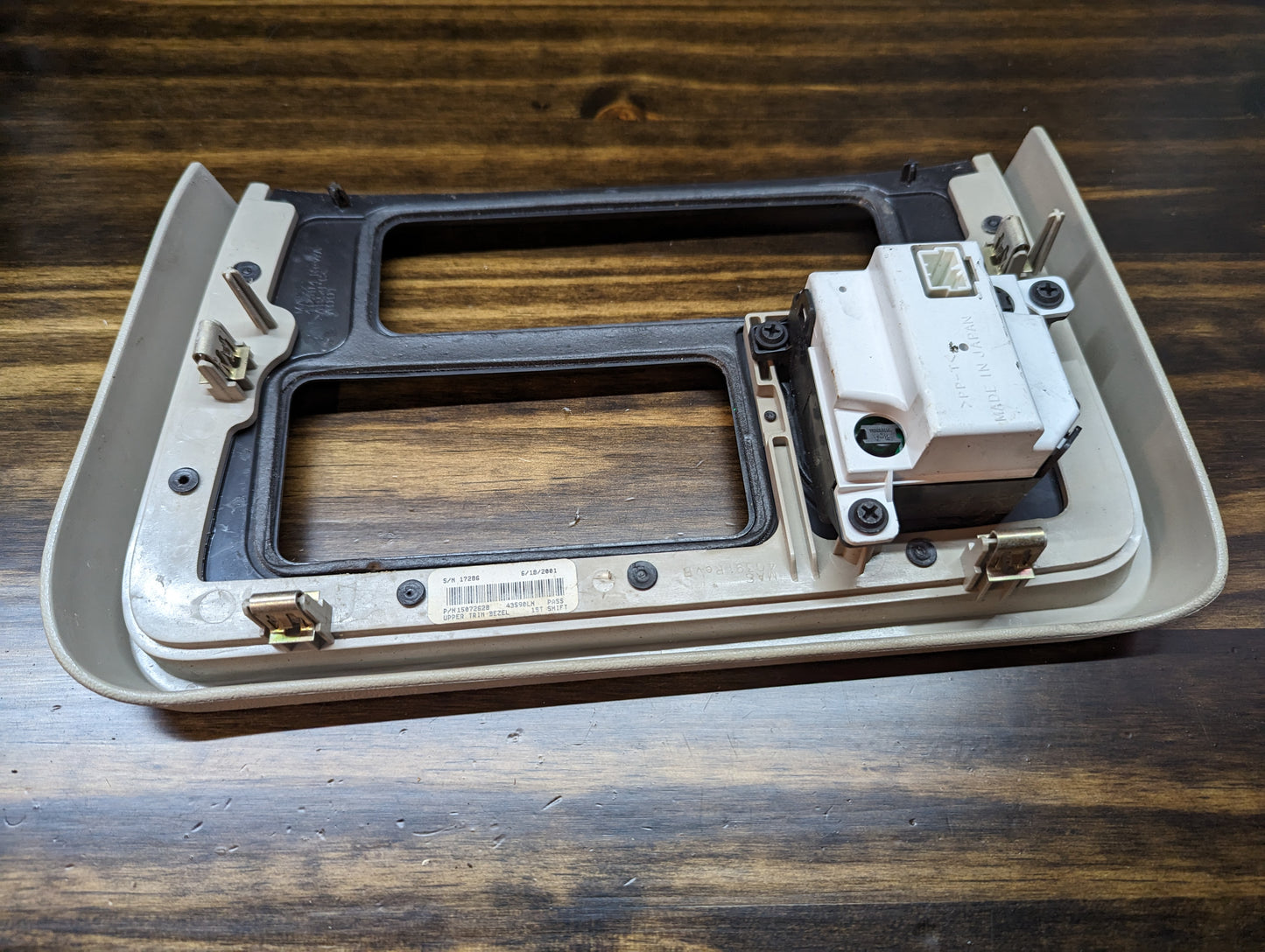 A General Motors OEM Center Console Front Bin Woodgrain Trim with Clock for 2002 Cadillac Escalade is shown on a wooden surface, featuring rectangular openings and an attached white electronic component.