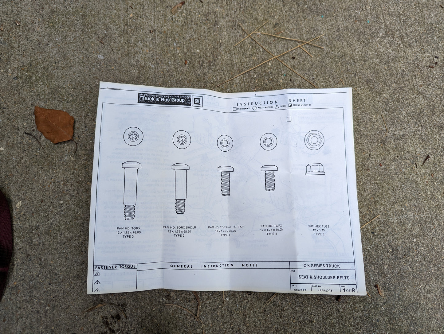 A printed instruction sheet for a General Motors NOS Passenger Seat Belt Retractor & Buckle in Red (1988-1994 Chevy/GMC C/K Trucks) lies on concrete, showing diagrams, labels, and torque specs for proper truck installation.