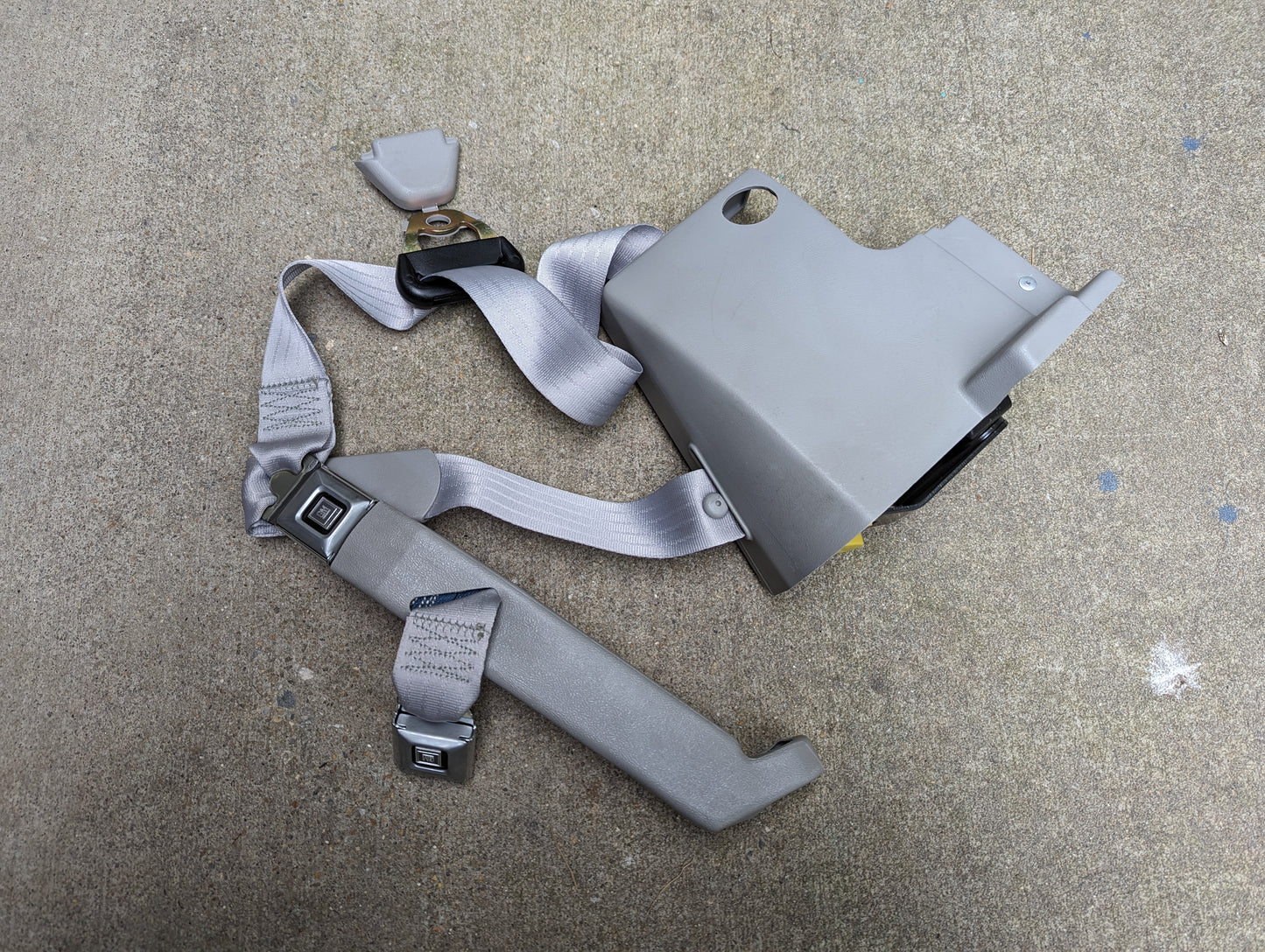 A gray NOS General Motors passenger seat belt retractor and buckle for 1982-1993 Chevy S10 or GMC S15, shown extended with plastic housing and mounting bracket on concrete. Suitable for restoring OEM safety.