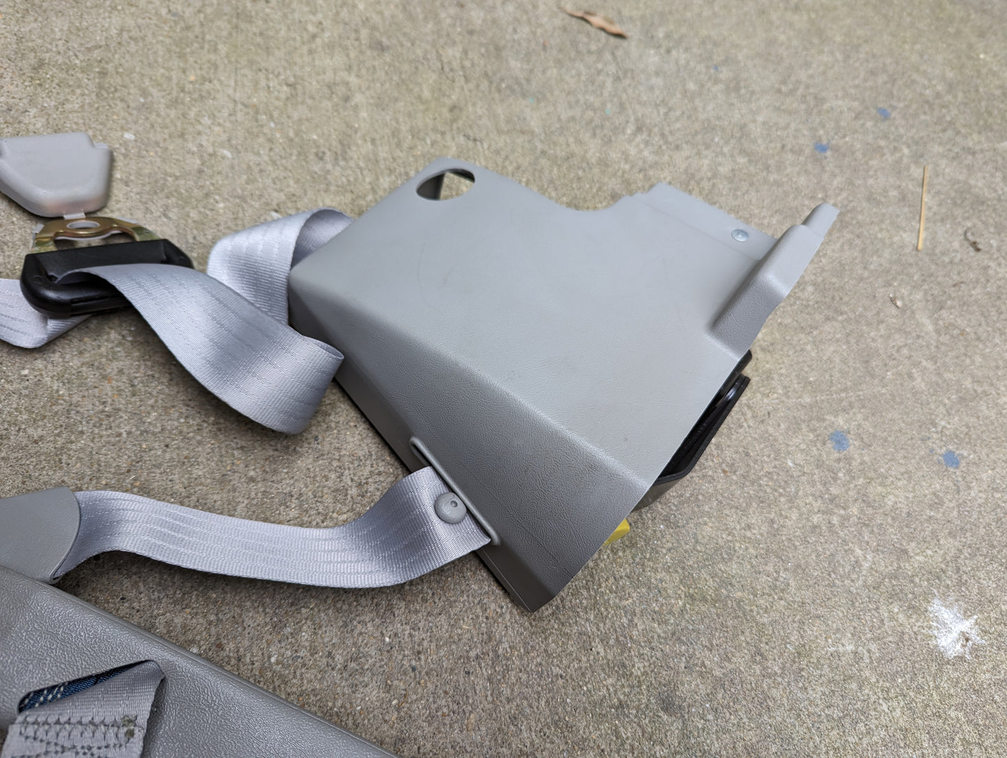 A General Motors NOS passenger seat belt retractor & buckle in gray for 1982-1993 Chevy S10/GMC S15 is lying on a concrete surface with its cover detached.