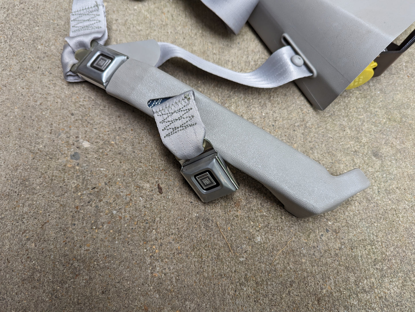 A close-up of a gray General Motors NOS Passenger Seat Belt Retractor & Buckle for 1982–1993 Chevy S10/GMC S15, with two square metal buckles and gray webbing, resting on concrete. Part of a plastic seat is visible in the upper right corner.