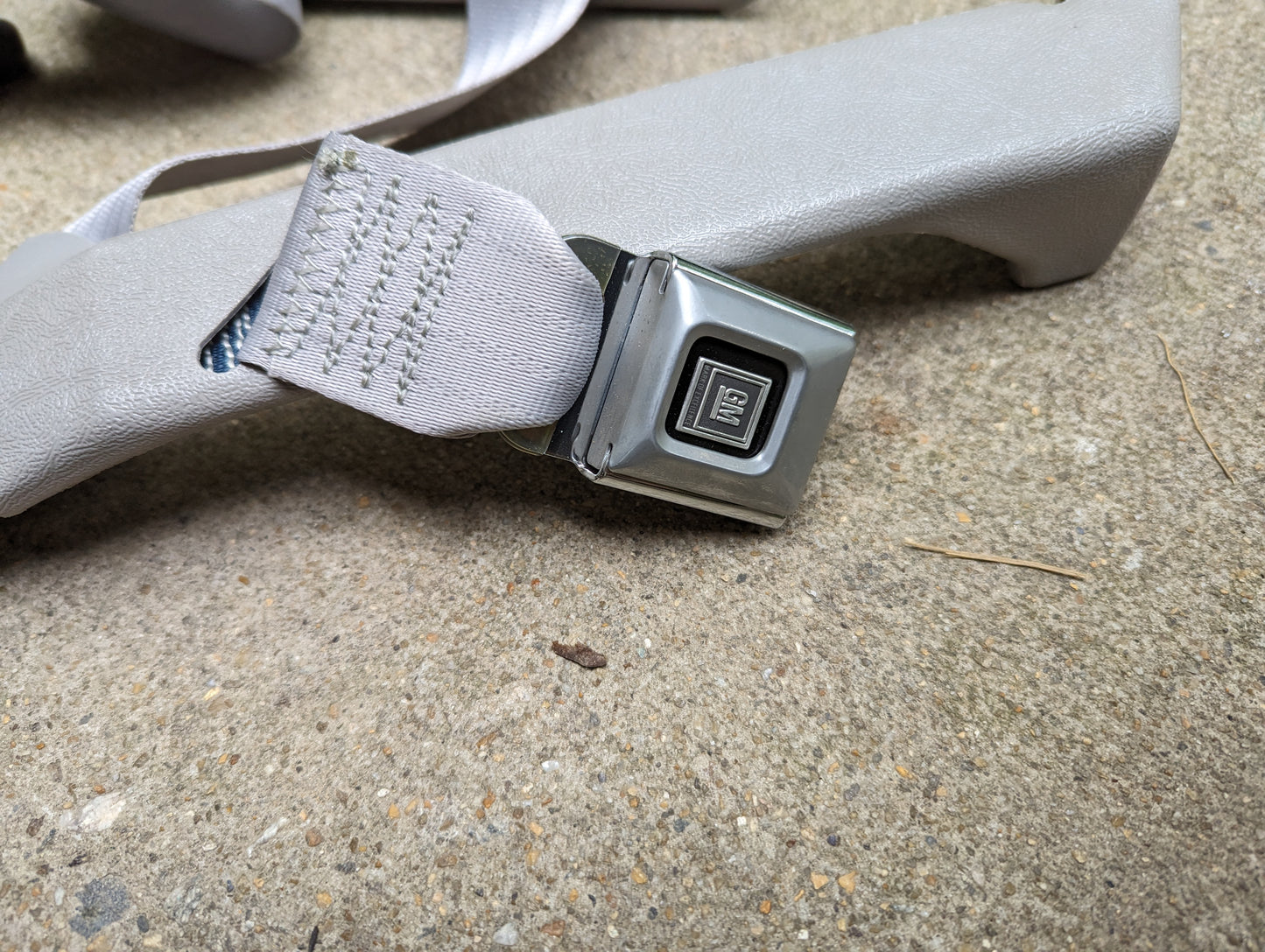 A close-up of a NOS gray passenger seat belt retractor and buckle for 1982-1993 Chevy S10 and GMC S15 by General Motors, resting on a textured concrete surface.
