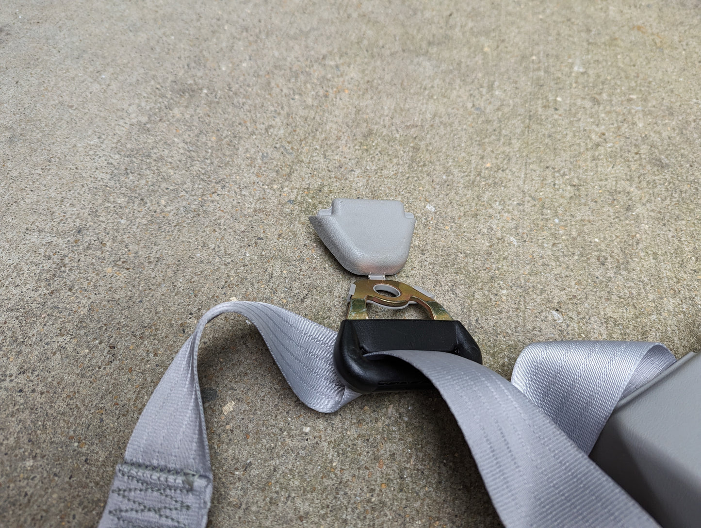 A close-up of a NOS General Motors gray passenger seat belt retractor and buckle for 1982-1993 Chevy S10/GMC S15, shown detached on a rough concrete surface.
