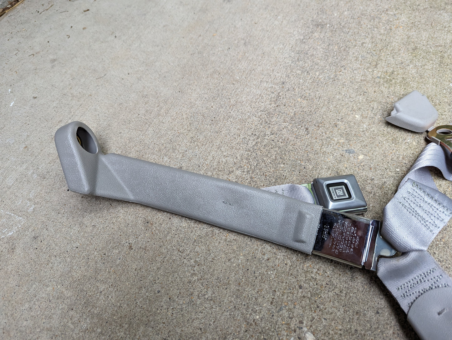 A General Motors NOS passenger seat belt retractor and buckle in gray for 1982-1993 Chevy S10/GMC S15 is shown lying on a concrete surface.