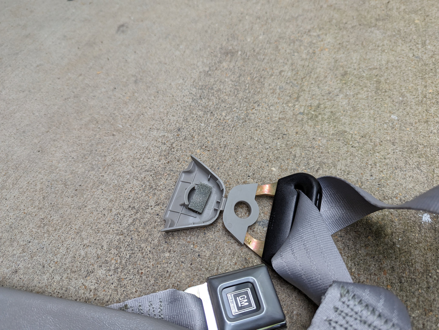 A close-up of a gray General Motors NOS passenger seat belt retractor and buckle for 1982-1993 Chevy S10/GMC S15 on concrete, with the seat belt and GM-labeled buckle partially visible.