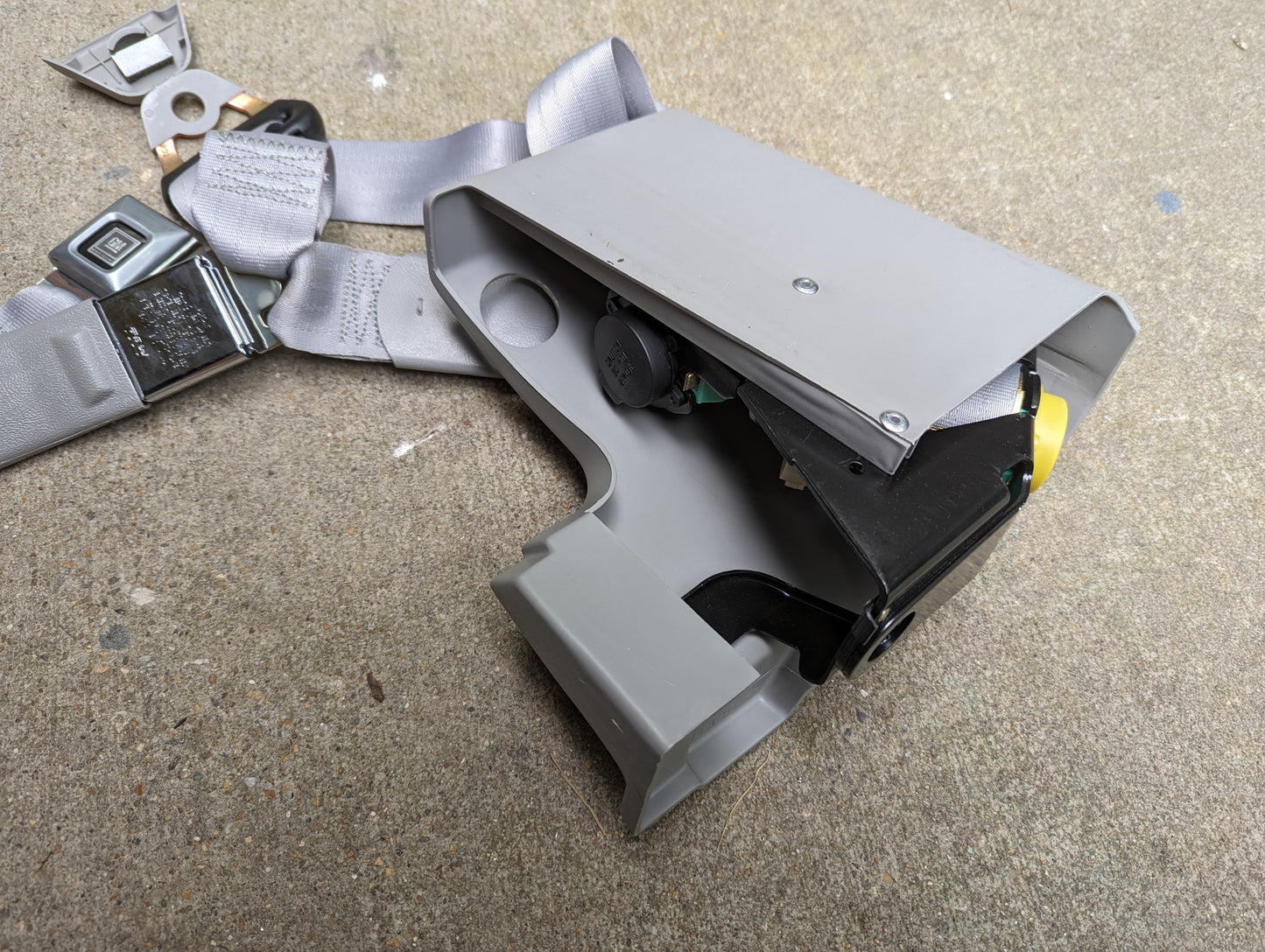 A NOS General Motors passenger seat belt retractor and buckle in gray for 1982–1993 Chevy S10/GMC S15 are shown disassembled on concrete, displaying internal parts and screws.