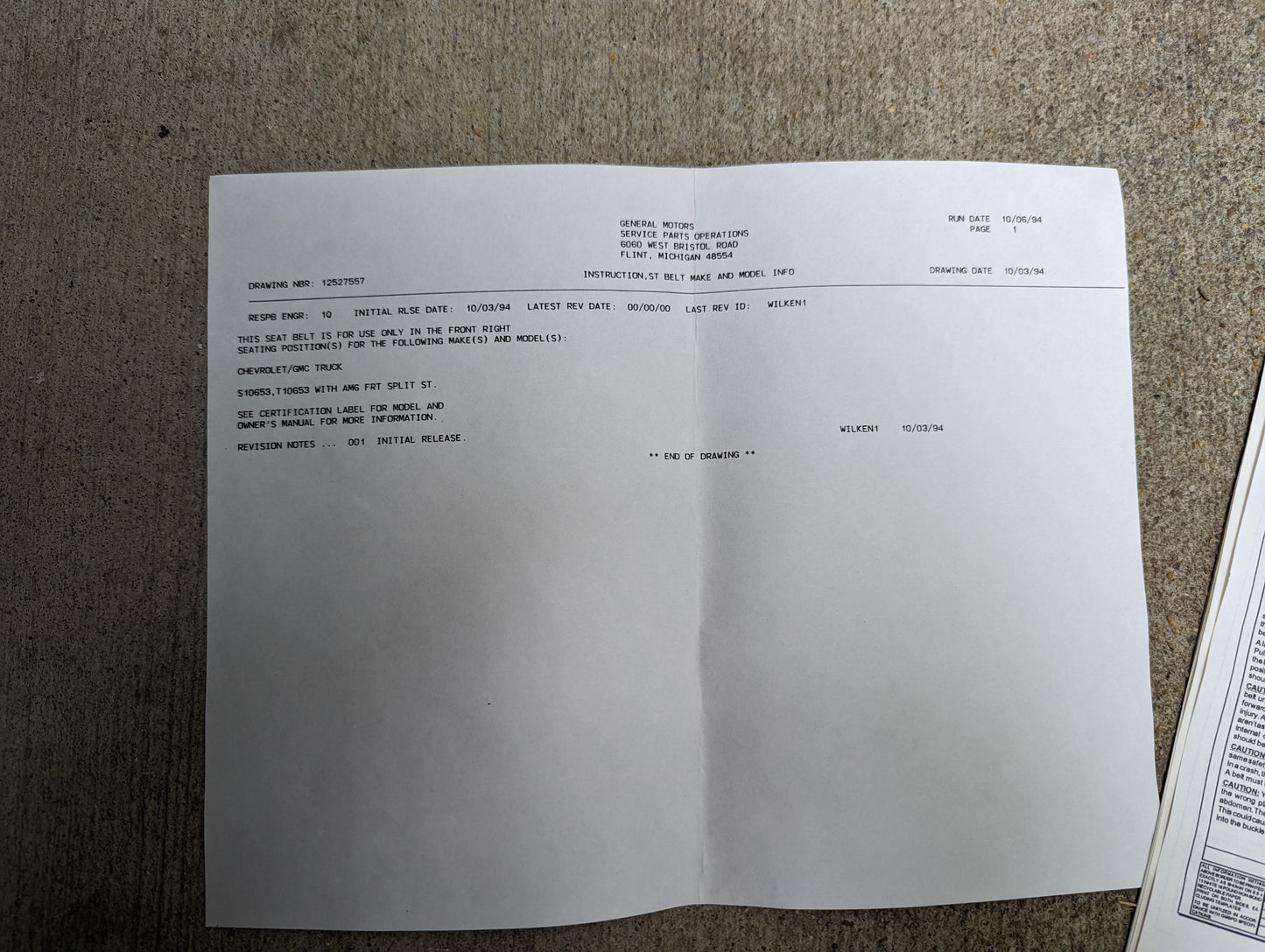 A printed document on white paper rests on a rough gray surface, showing codes, descriptions, and dates—possibly referencing a General Motors NOS passenger seat belt retractor & buckle in gray for 1982-1993 Chevy S10/GMC S15. Another paper peeks at the edge.