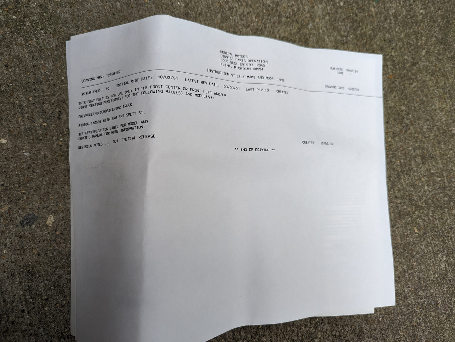 Close-up of a printed document on concrete, displaying official discharge papers for a NOS Center Seat Belt & Buckle in Beige (1982-1993 Chevy S10, GMC S15) by General Motors. The paper is slightly curved and shadowed at the edges.