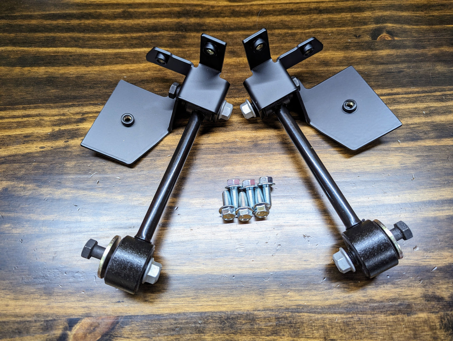 Two black metal brackets with rods and bushings from The Ugly Truckling KIT ZQ8/Xtreme Rear Sway Bar for 1982-2004 Chevy S10/GMC S15/Sonoma are laid out on wood, with bolts and nuts arranged between them.