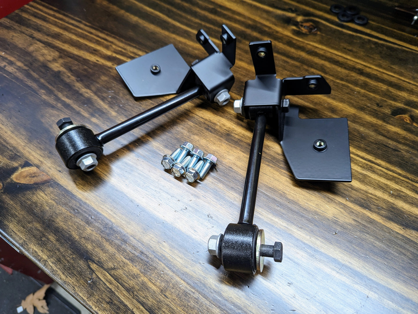Two black metal brackets with bushings and bolts from The Ugly Trucklings KIT ZQ8/Xtreme Rear Sway Bar for 1982-2004 Chevrolet S10, GMC S15, Sonoma are shown on a polished wood surface with several loose screws between them.