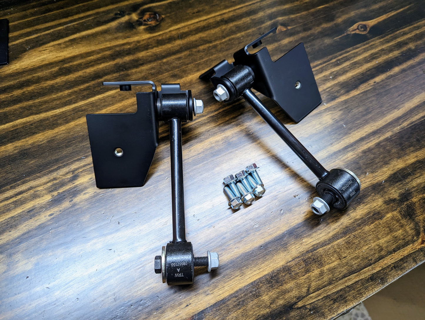 Two black metal suspension components from The Ugly Truckling KIT ZQ8/Xtreme Rear Sway Bar with NEW Brackets for 1982-2004 Chevrolet S10, GMC S15, Sonoma rest on wood, with included silver bolts and nuts between them.