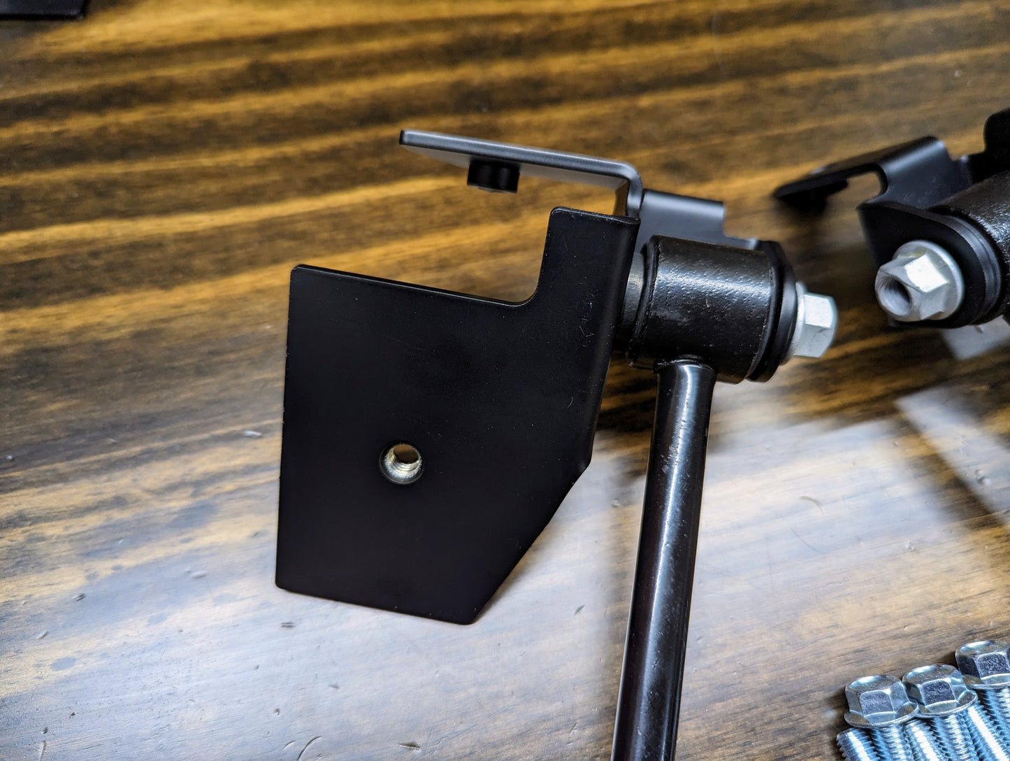 Close-up of a black metal bracket with bolt from The Ugly Truckling KIT ZQ8/Xtreme Rear Sway Bar with NEW Brackets for 1982-2004 Chevrolet S10, GMC S15, Sonoma—essential for your sway bar upgrade. Screws and bolts are also shown on a wooden surface.