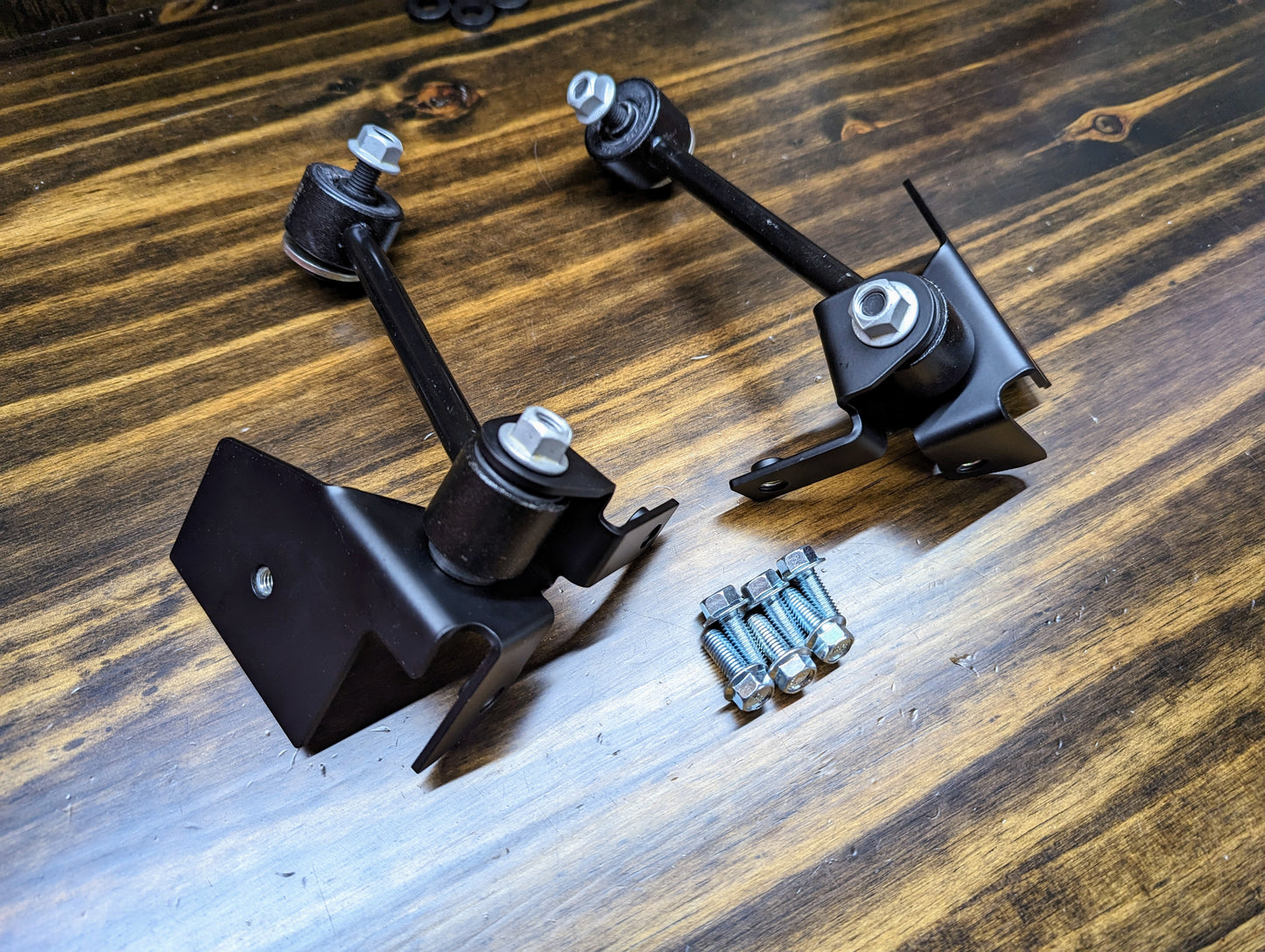 Two black brackets with bushings and bolts from The Ugly Truckling KIT ZQ8/Xtreme Rear Sway Bar for 1982-2004 Chevrolet S10, GMC S15, Sonoma are on a wooden surface, with four small silver bolts between the brackets.
