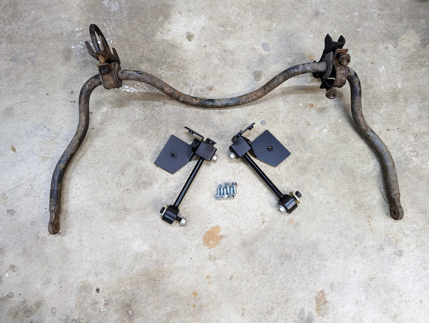 A rusty Chevrolet S10 sway bar sits on concrete beside new black end links, brackets, and bolts—ready for install with The Ugly Truckling KIT ZQ8/Xtreme Rear Sway Bar & NEW Brackets for 1982-2004 Chevy S10/GMC S15/Sonoma.