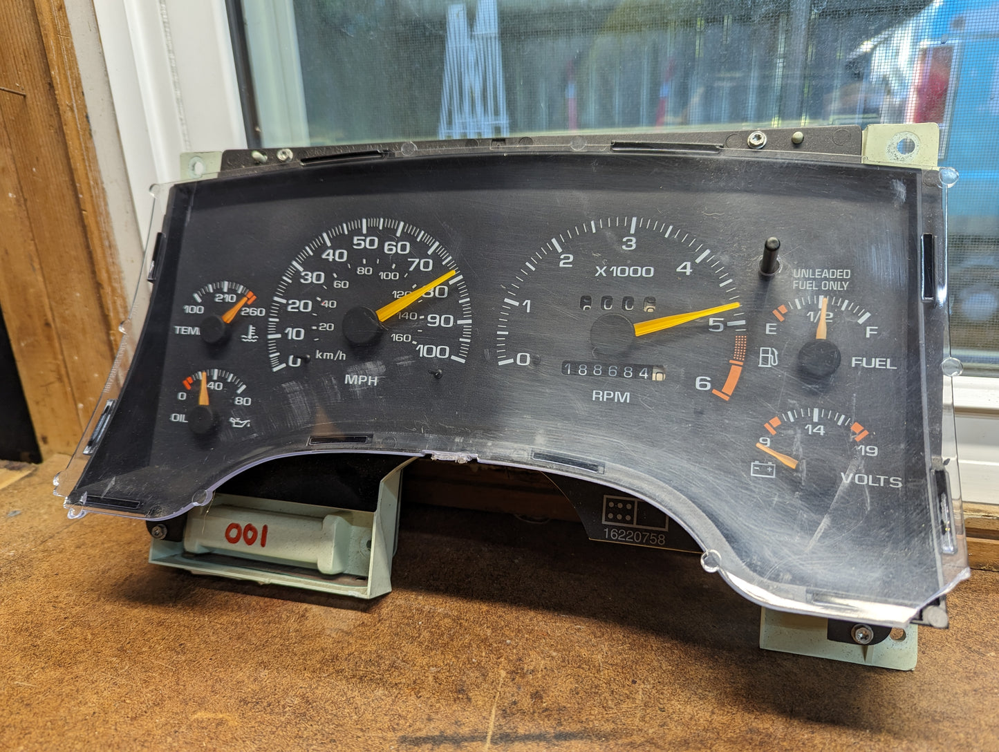 A General Motors OEM Instrument Cluster with Tachometer for 1995-1997 Chevy S10 or Blazer with Console Shifter is shown close-up on a wooden surface near a window, displaying speed, RPM, temp, fuel, oil pressure, and volts gauges.