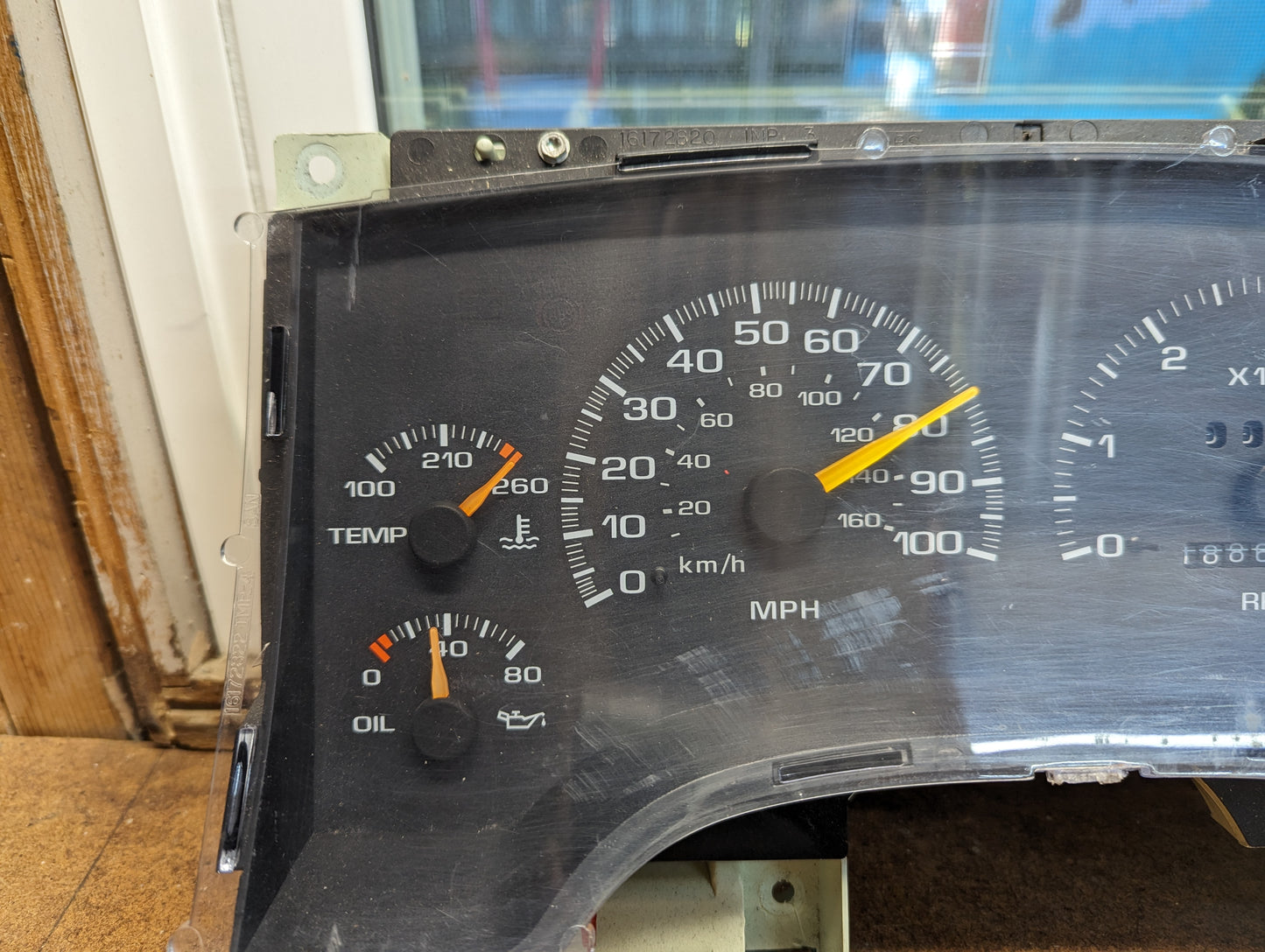 A close-up of the OEM Instrument Cluster with Tachometer by General Motors for 1995-1997 Chevy S10 or Blazer (with console shifter) shows white-marked gauges and orange needles; the dashboard is removed from the car.