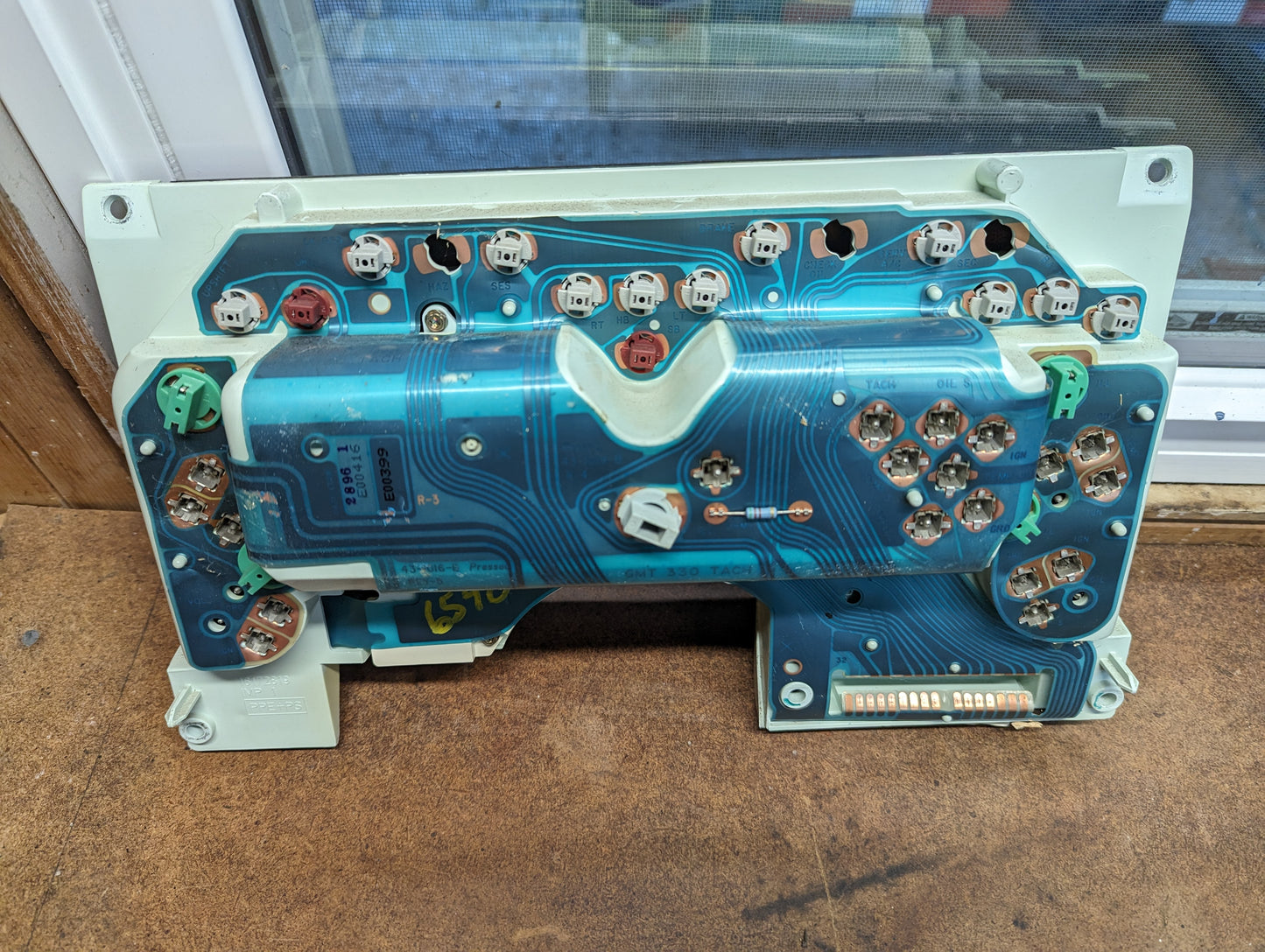 A General Motors OEM Instrument Cluster with Tachometer for 1995-1997 Chevy S10 or Blazer with Console Shifter, showing its blue circuit board, connectors, bulbs, and components on a brown surface near a window.
