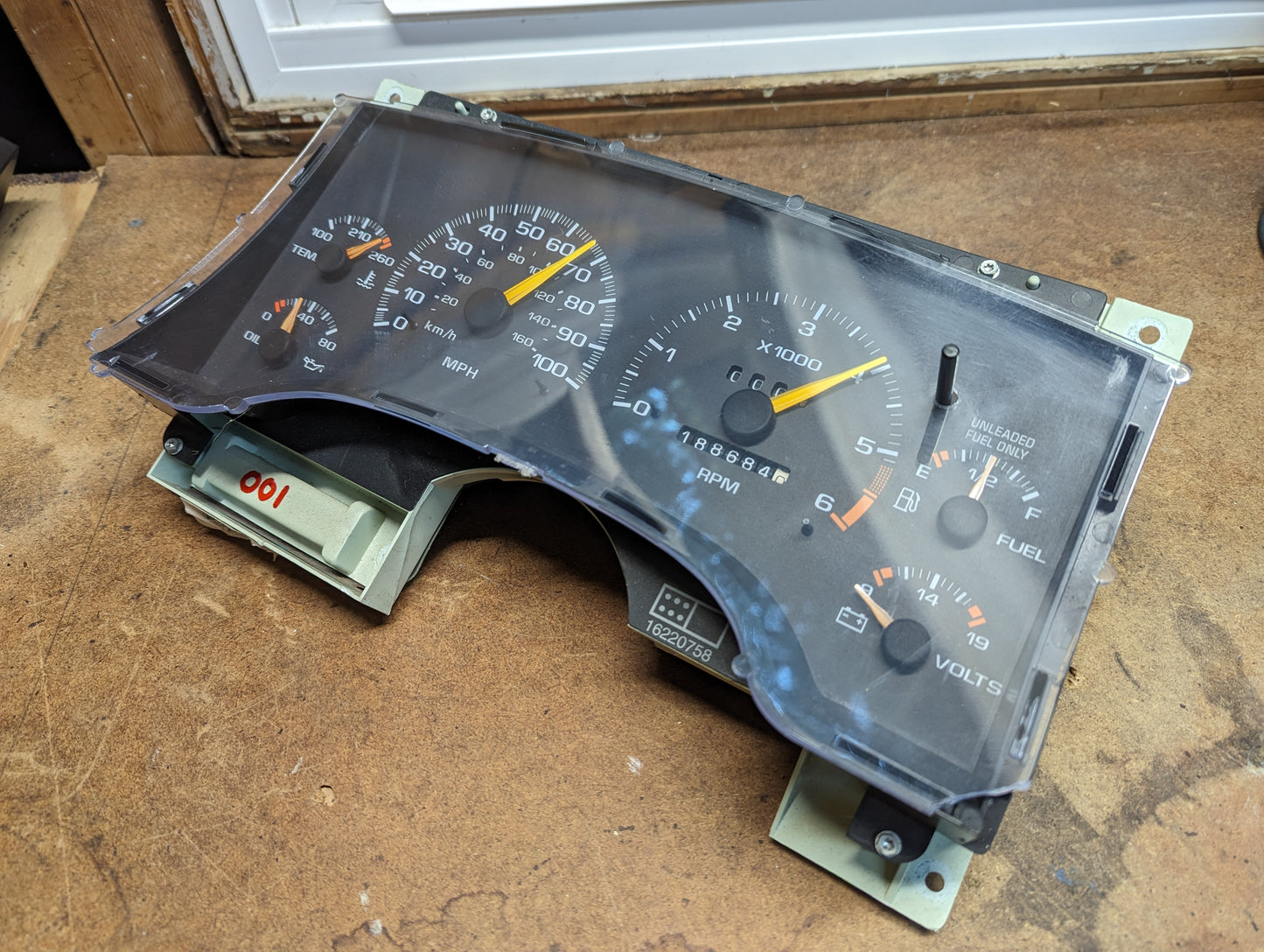 A General Motors OEM Instrument Cluster with Tachometer for 1995-1997 Chevy S10 or Blazer (with Console Shifter and more) is shown close-up, displaying speedometer, fuel, oil, voltage, and temperature gauges on a wooden surface near a window.
