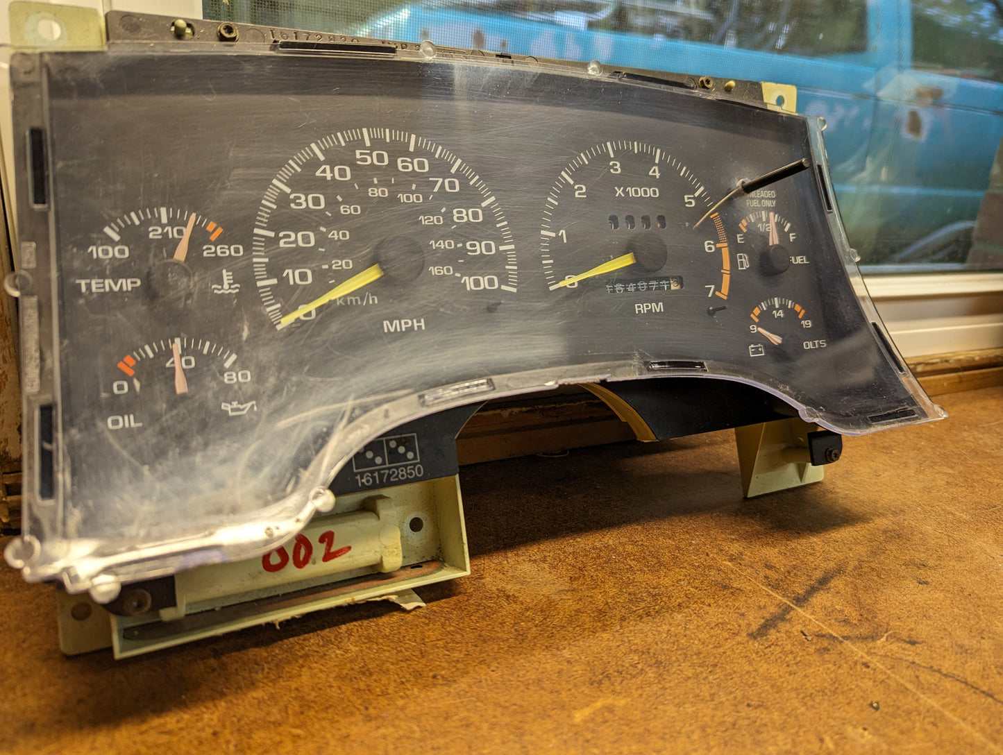 A removed General Motors OEM Instrument Cluster with Tachometer for 1995-1997 Chevy S10 or Blazer with Console Shifter rests on a wooden surface, showing analog gauges. A window and a blue vehicle are visible in the background.